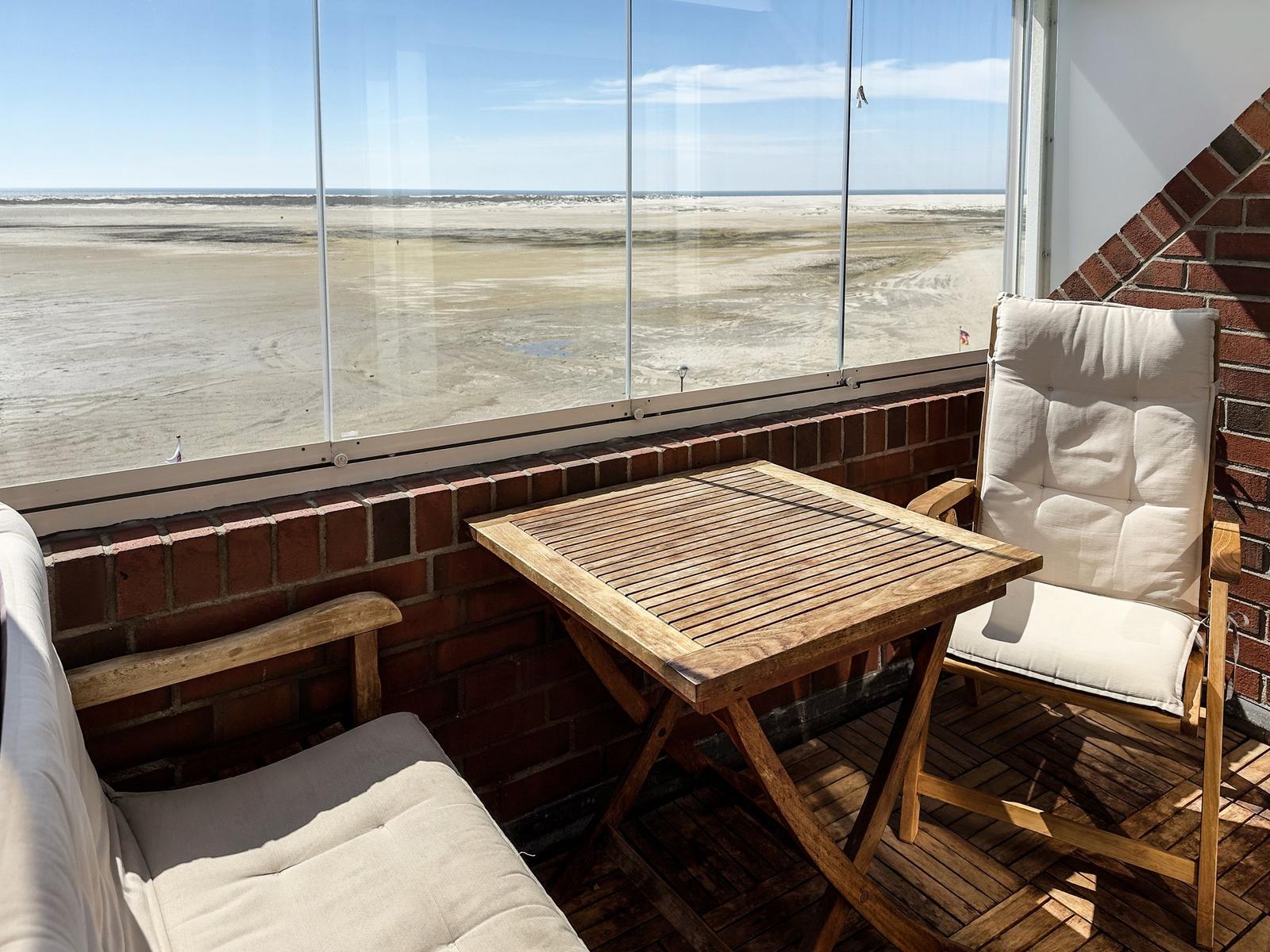 Terrasse mit Tisch und Stühlen. Blick auf Strand und Meer durch Glaswand.