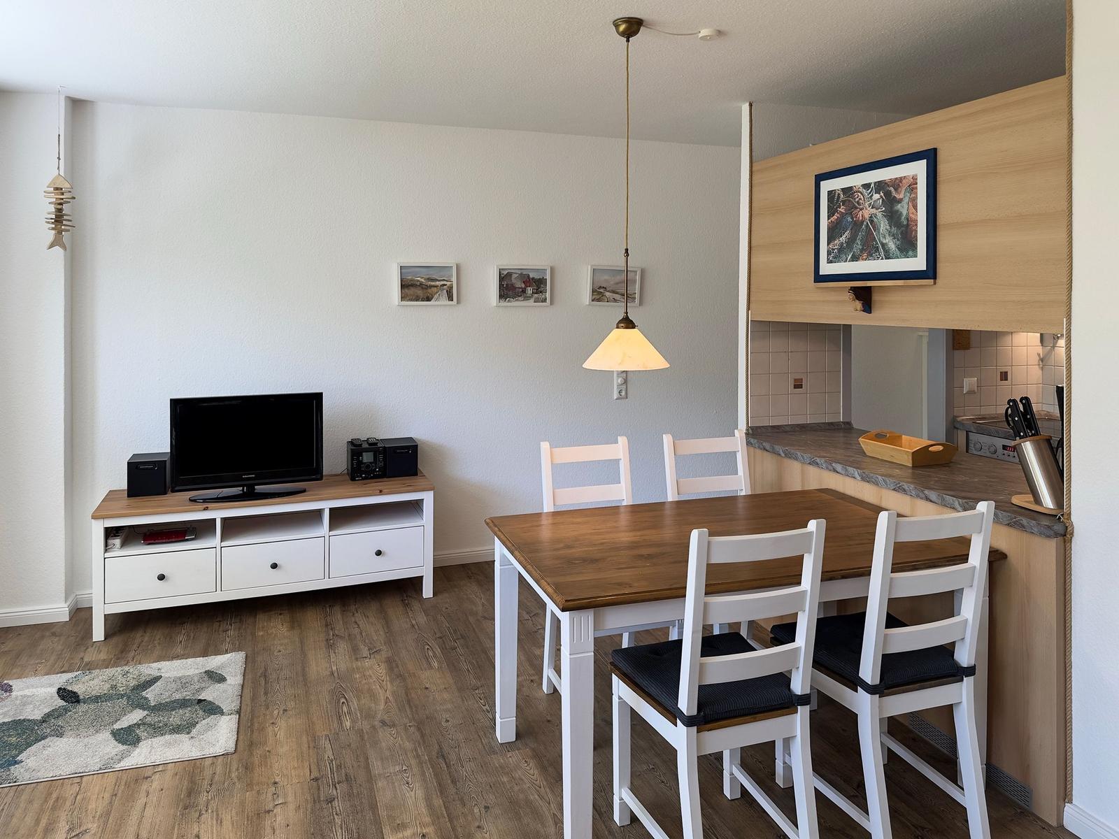 Essbereich mit Tisch und Stühlen, Küche und Wohnbereich mit Fernseher und Wandbildern.