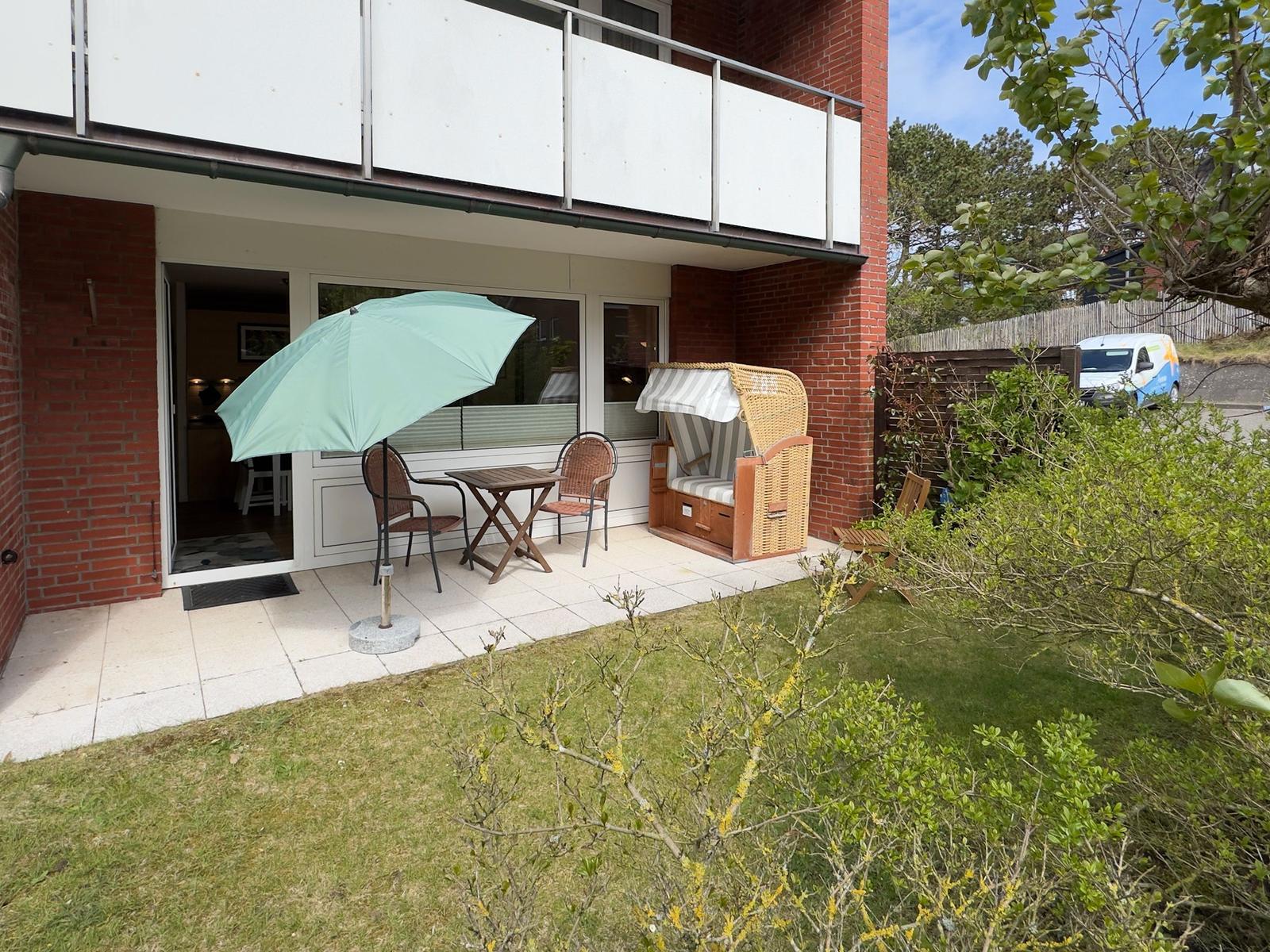 Terrasse mit Tisch, Stühlen und Sonnenschirm. Strandkorb und Grünfläche im Hintergrund.