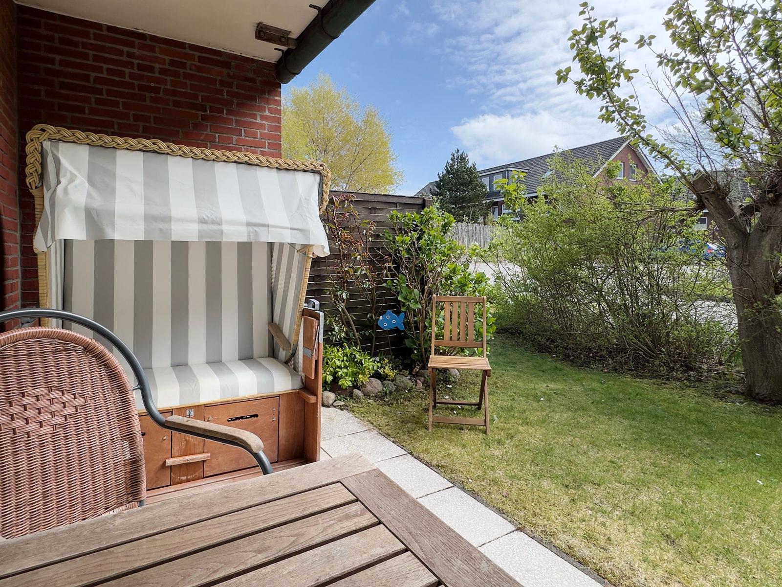 Terrasse mit Liege, Tisch und Stuhl im Garten. Hintergrund: Gras, Bäume und Haus.