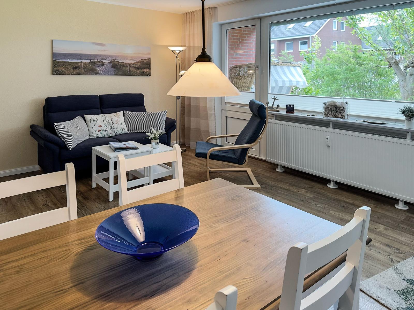 Wohn- und Essbereich mit Sofa, Stuhl und Tisch. Große Fenster mit Blick nach draußen.