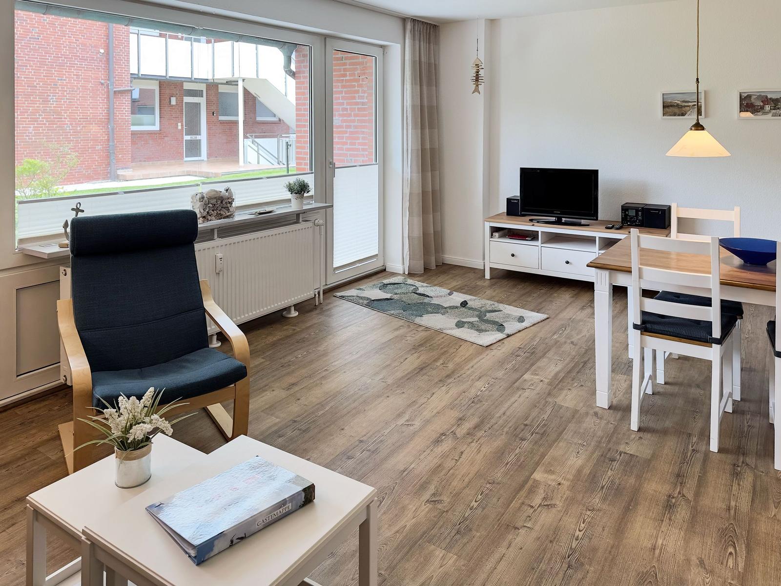 Wohn- und Essbereich mit Holzboden, TV, Esstisch und Sessel. Große Fensterfront mit Blick auf Außenbereich.
