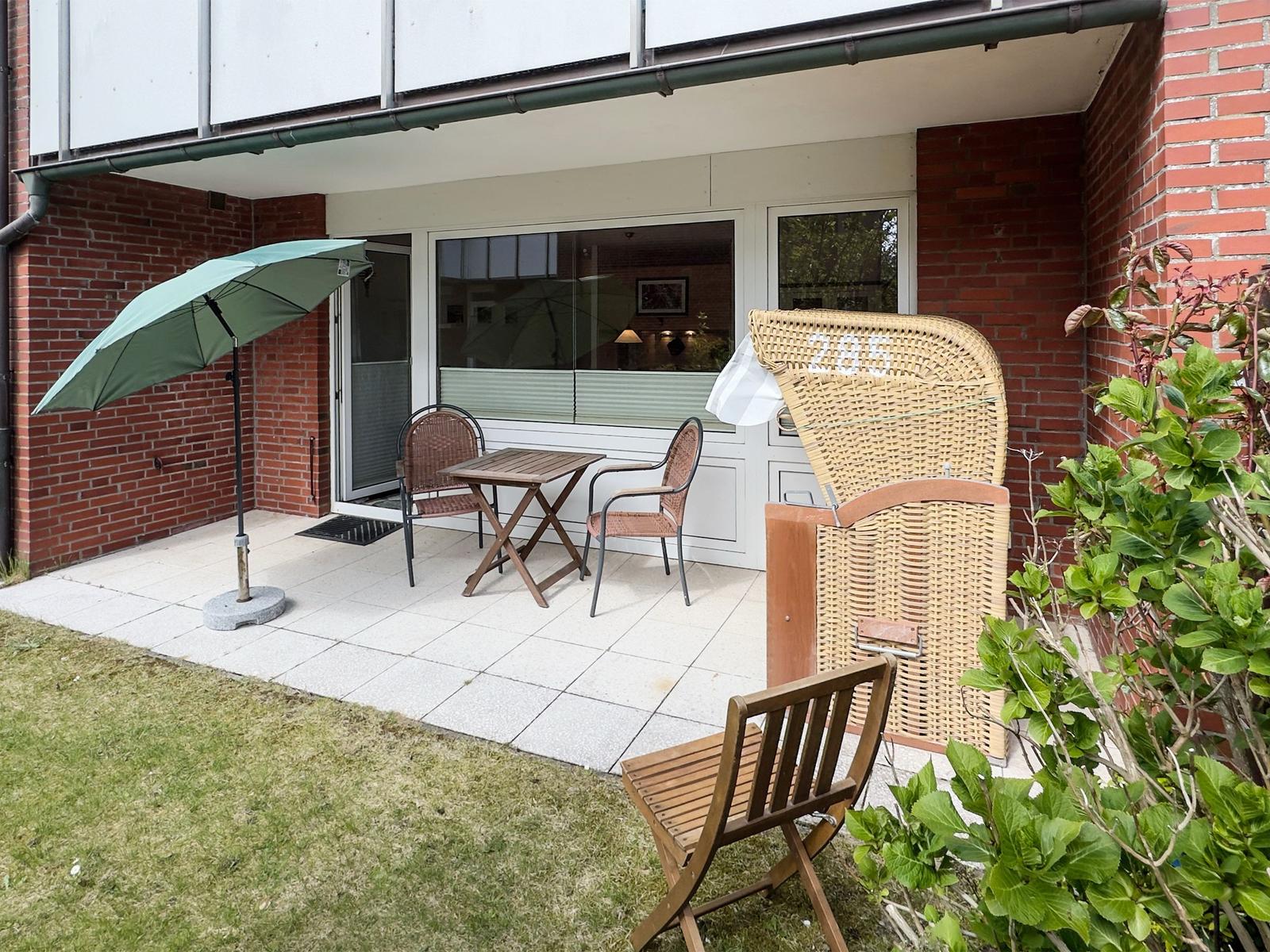 Terrasse mit Tisch, zwei Stühlen und Sonnenschirm. Holzstuhl und Korb neben grüner Pflanze.