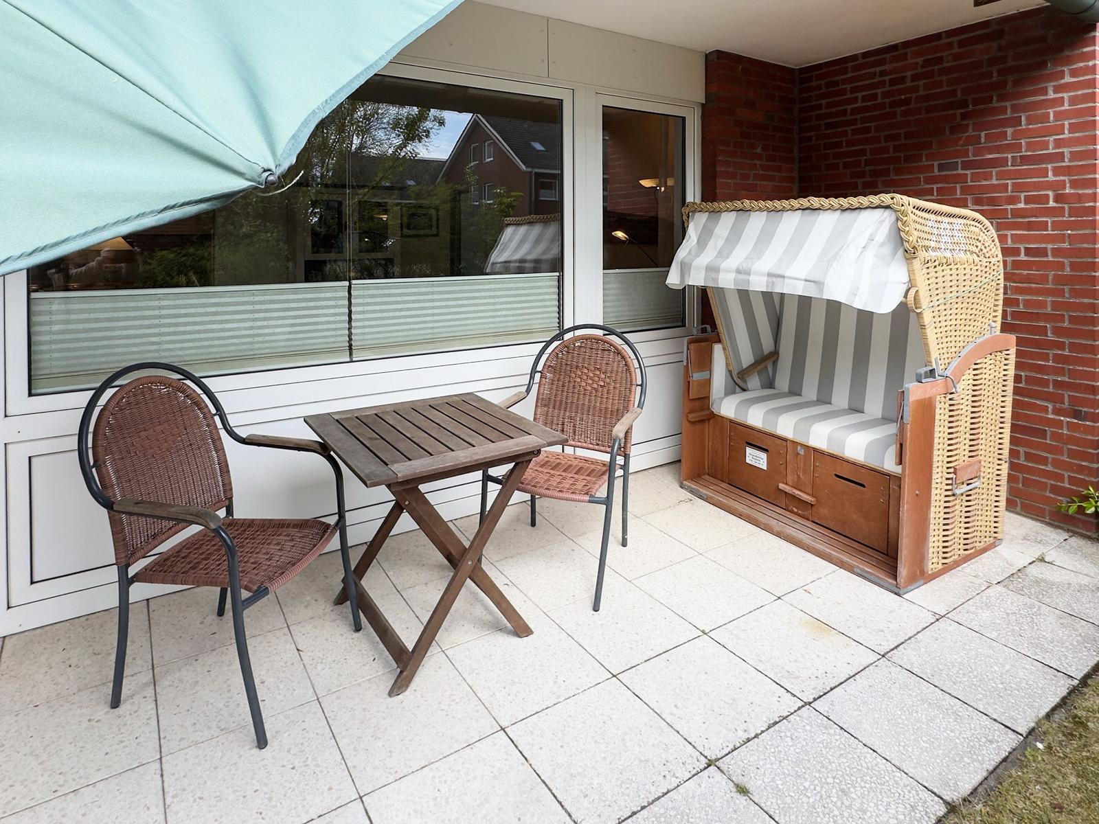 Terrasse mit Tisch, zwei Stühlen und Strandkorb. Hintergrund: Glasfront und Backsteinmauer.