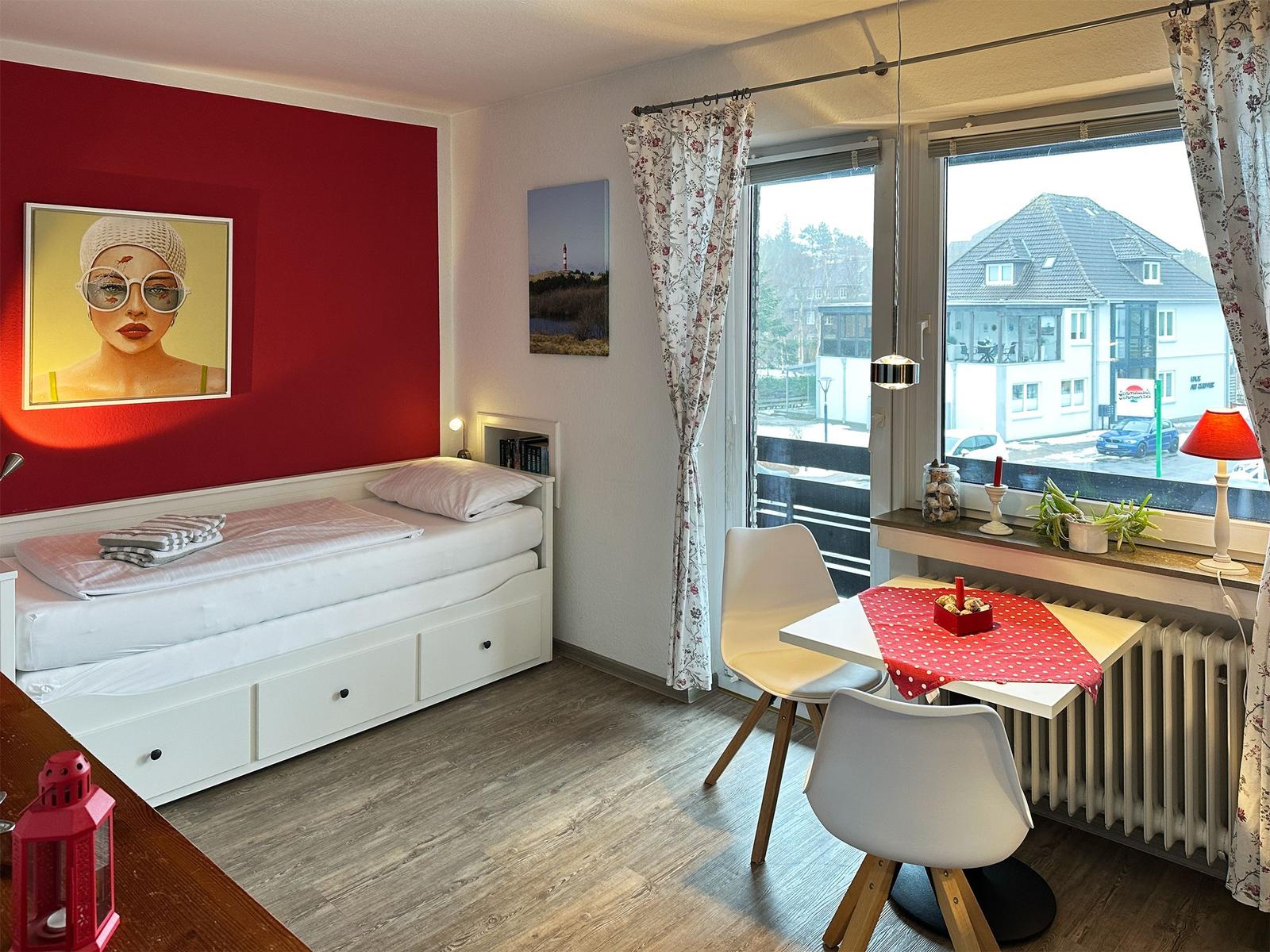 Schlafzimmer mit weißem Bett und roter Wand. Tisch mit Stühlen und Fensterblick.