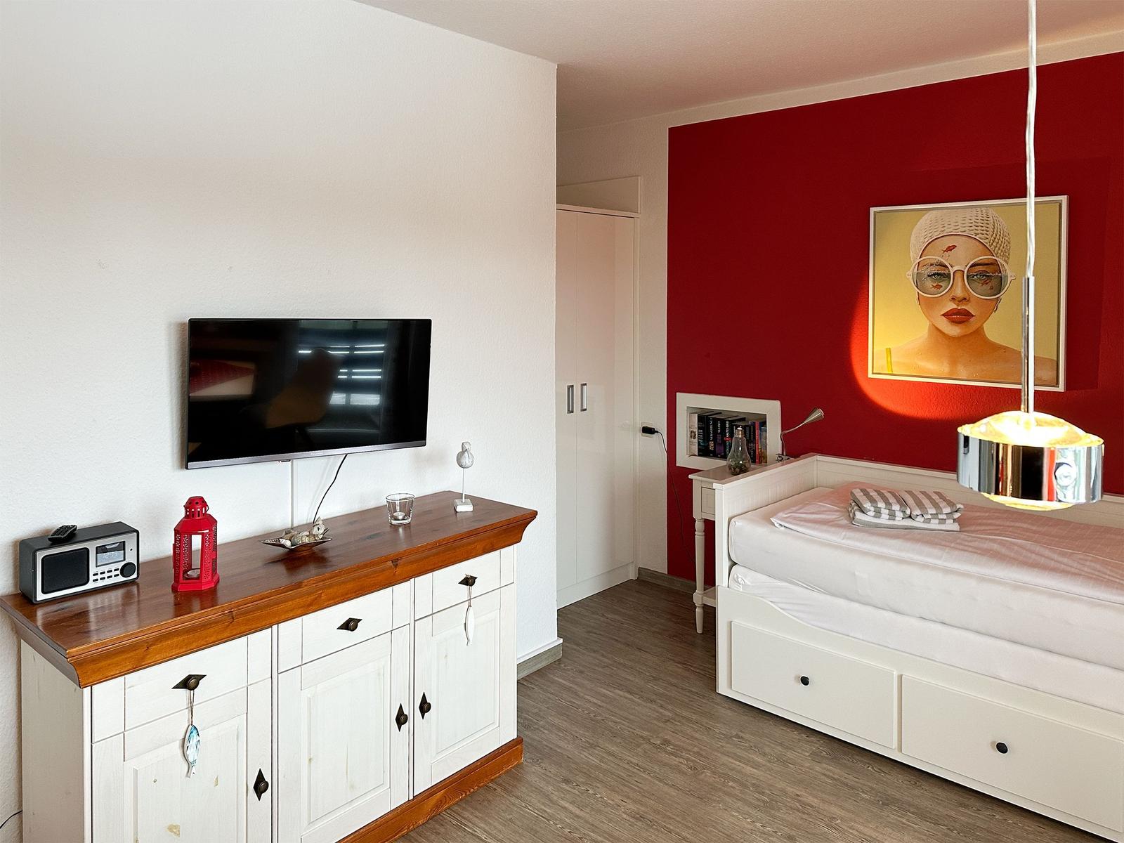 Schlafzimmer mit weißem Bett, roter Wand und Fernseher auf Holzschrank.