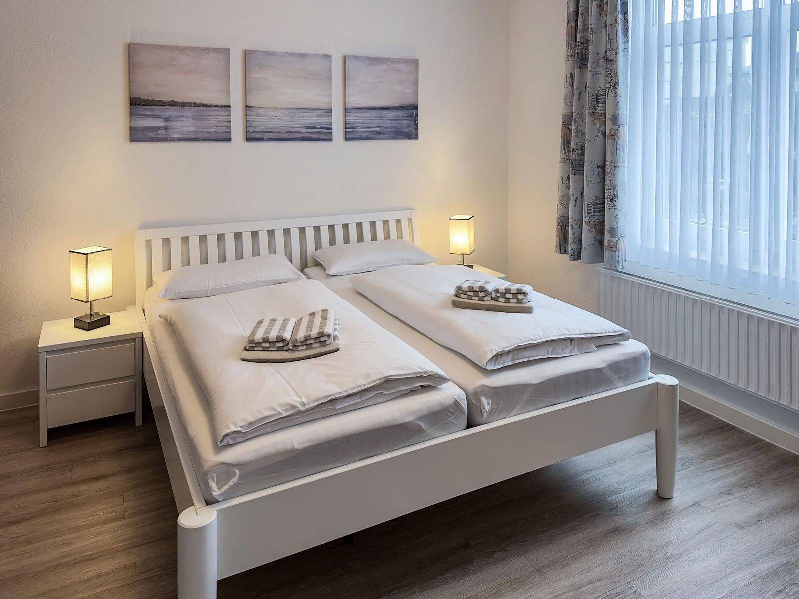 Ein Schlafzimmer mit weißem Bett, zwei Nachttischen und einem Fenster mit Vorhängen.