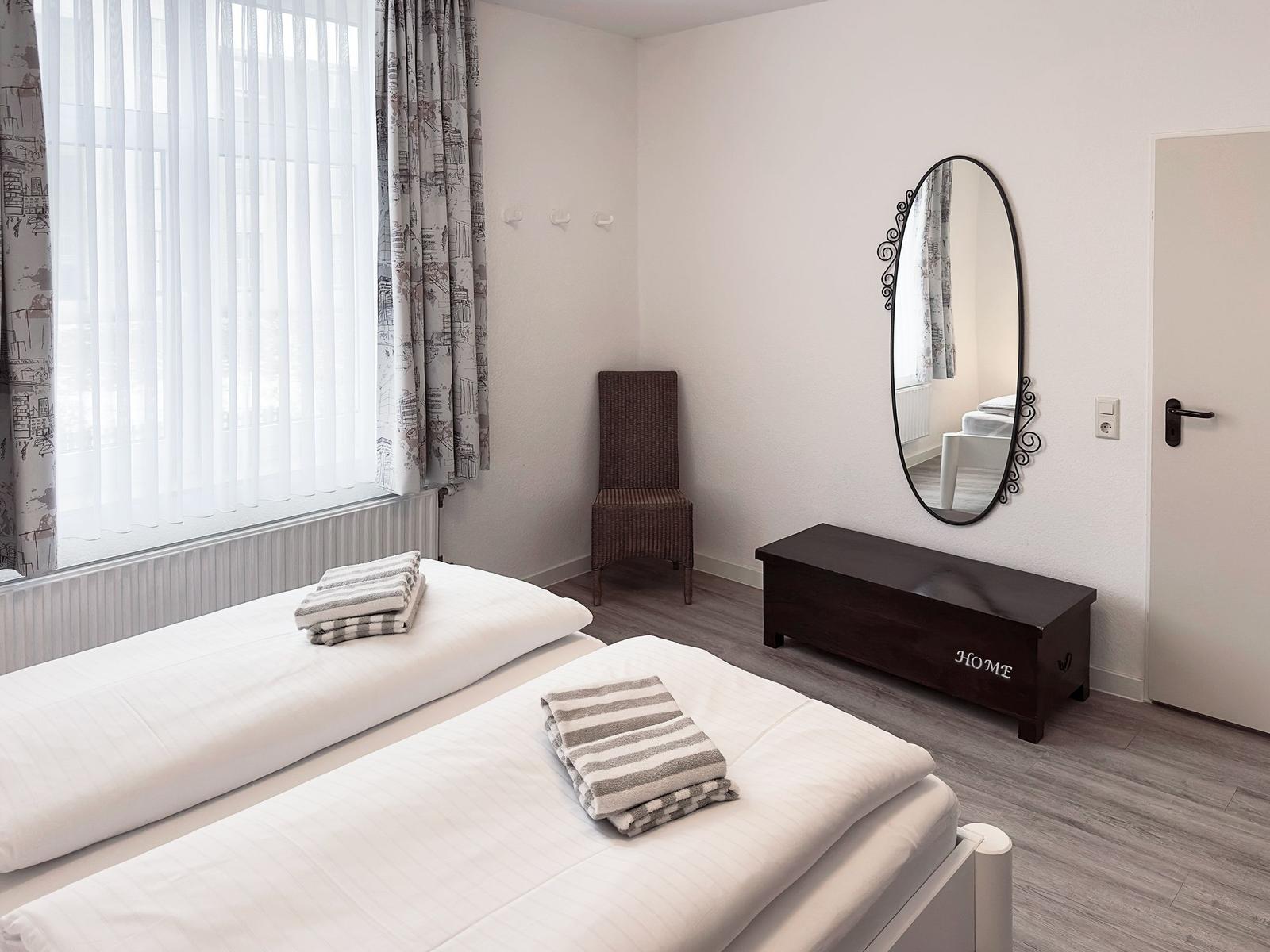 Zimmer mit zwei Betten, Spiegel, Stuhl und Fenster mit Vorhängen.