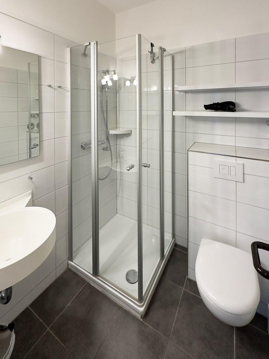 Badezimmer mit Dusche, Waschbecken und WC. Weiße Fliesen und graue Bodenfliesen.