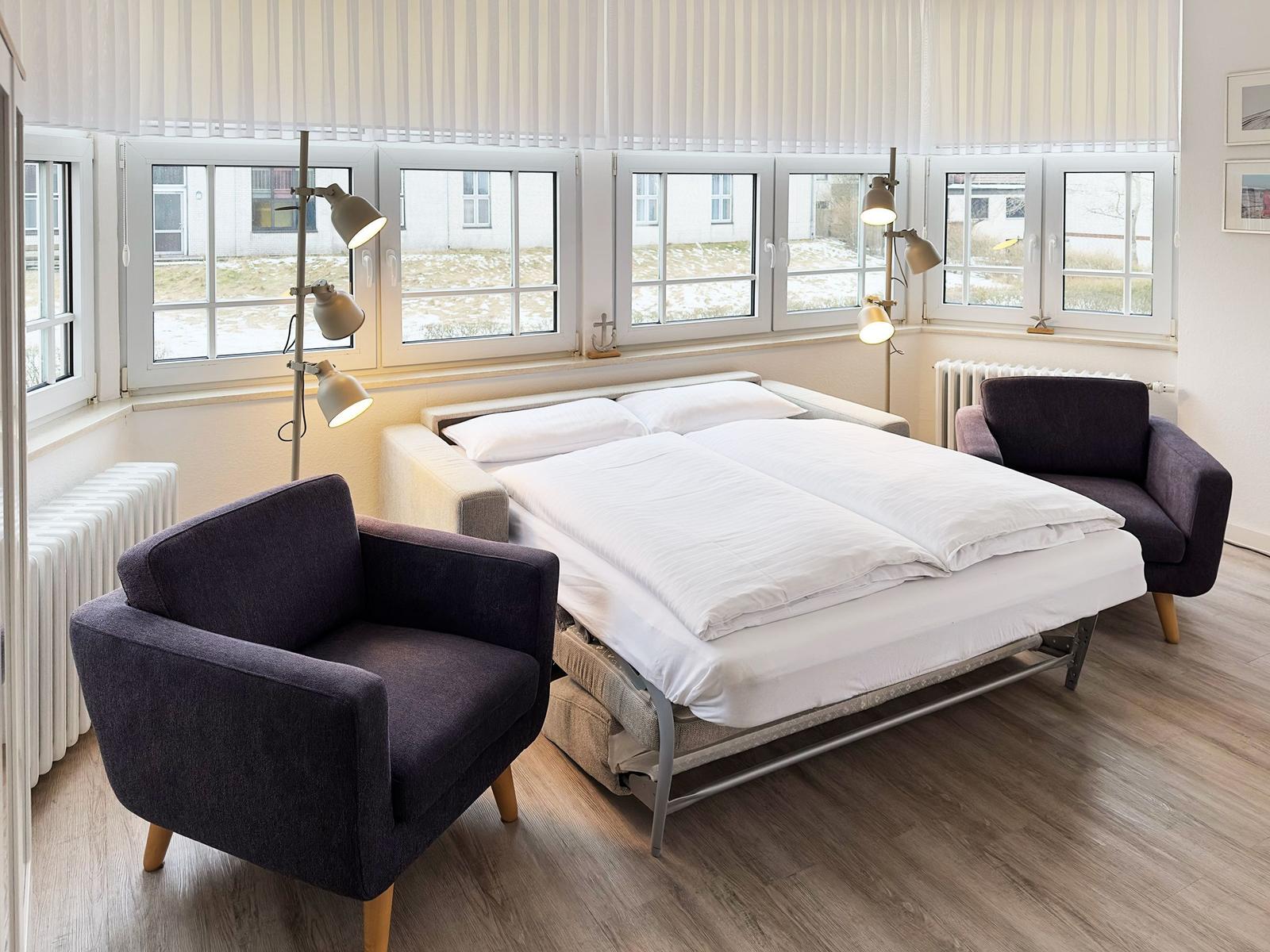 Schlafzimmer mit Bett, zwei Sesseln und Fenstern mit Blick nach draußen.