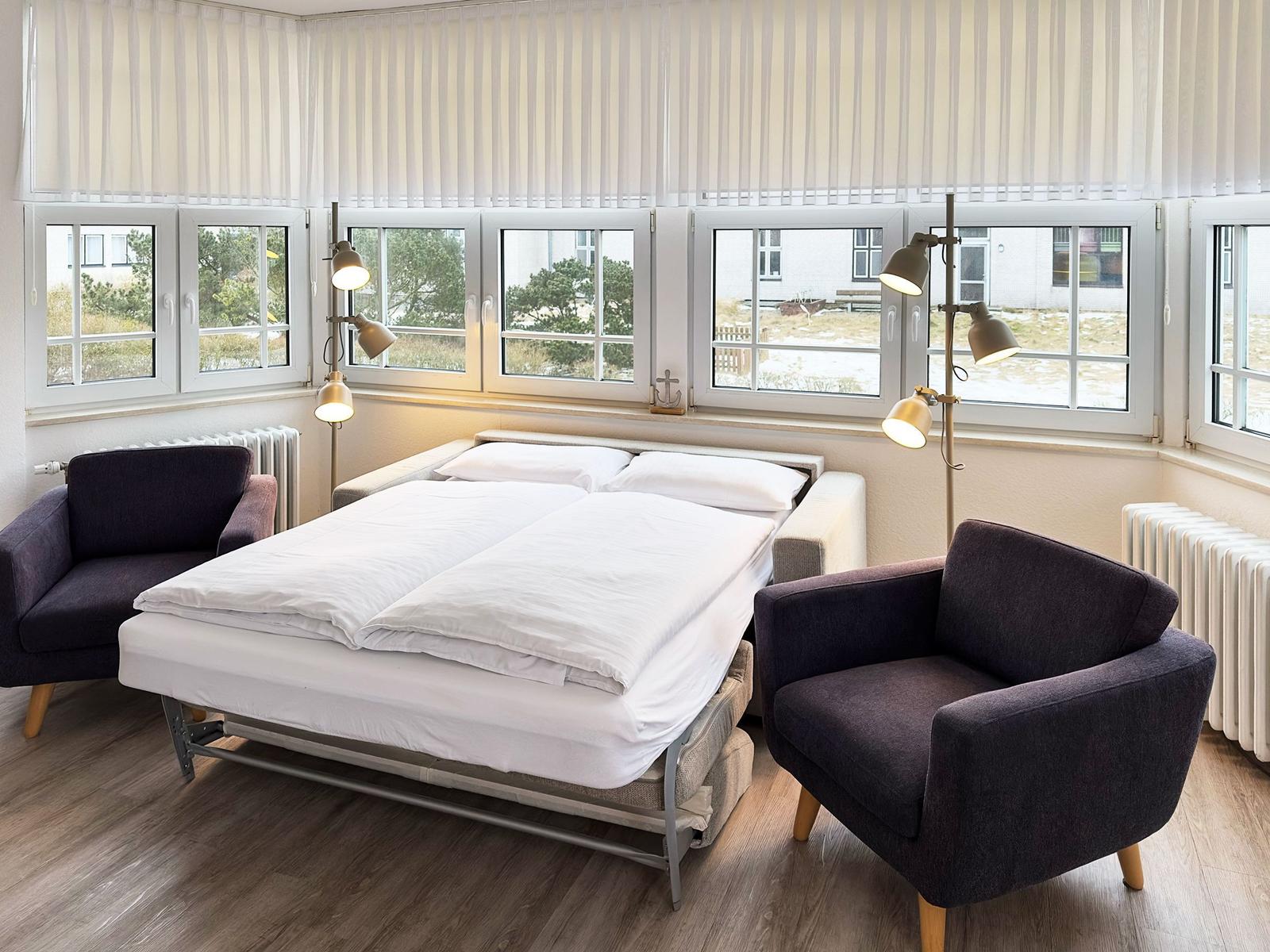 Schlafzimmer mit Bett, zwei Sesseln und Fenstern mit Jalousien.