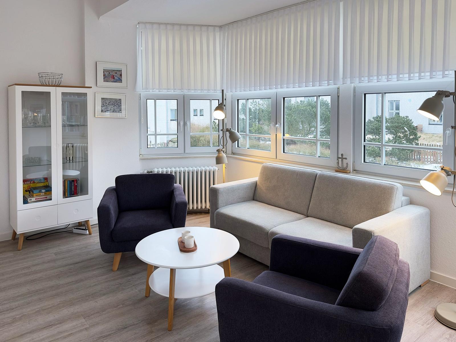 Wohnzimmer mit Sofa, zwei Sesseln, Tisch und Fenster. Regal und Lampen sind vorhanden.
