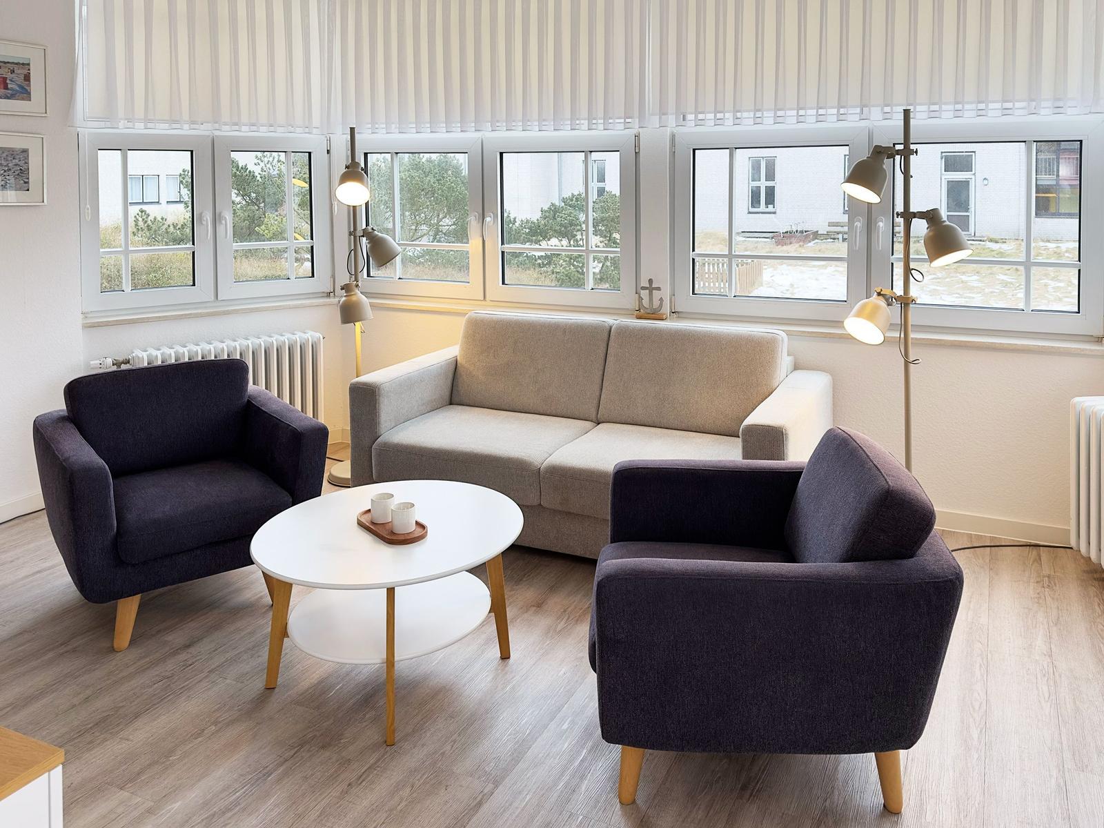 Wohnzimmer mit Sofa und zwei Sesseln, runder Tisch und Fenster mit Blick nach draußen.