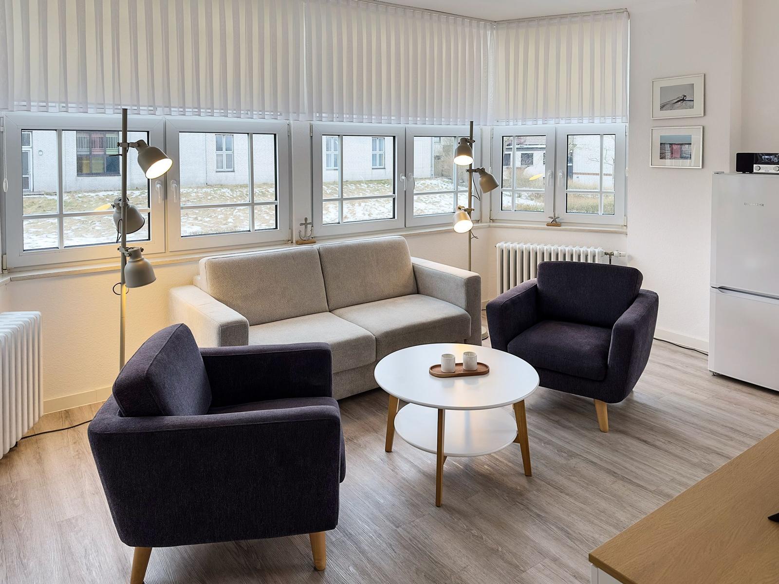 Wohnzimmer mit Sofa, zwei Sesseln und runden Tisch. Fenster mit Jalousien und Kühlschrank.