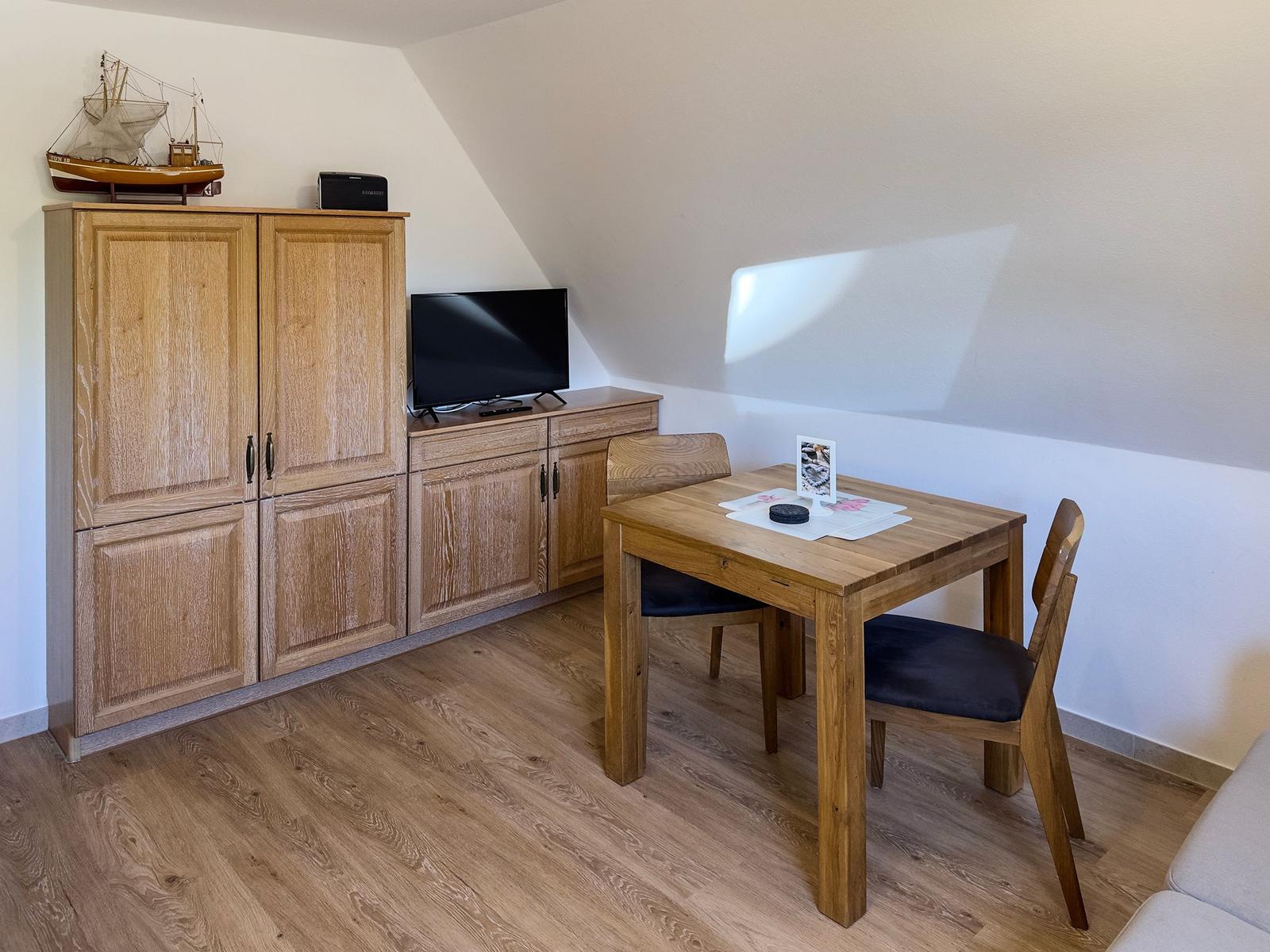 Holztisch mit zwei Stühlen, Fernseher und Schrank unter schrägem Dach.