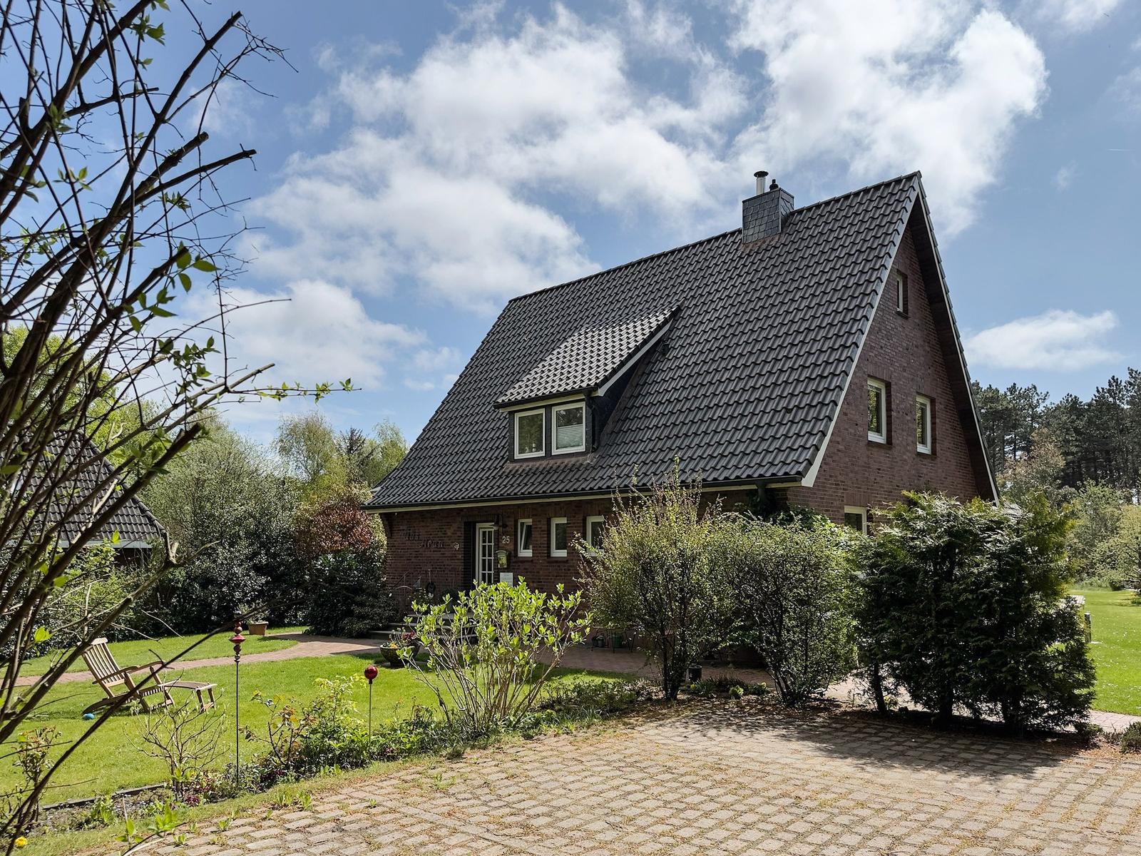 Ein zweistöckiges Haus mit dunklem Dach und rotem Backstein, umgeben von Grün und einem gepflasterten Weg.