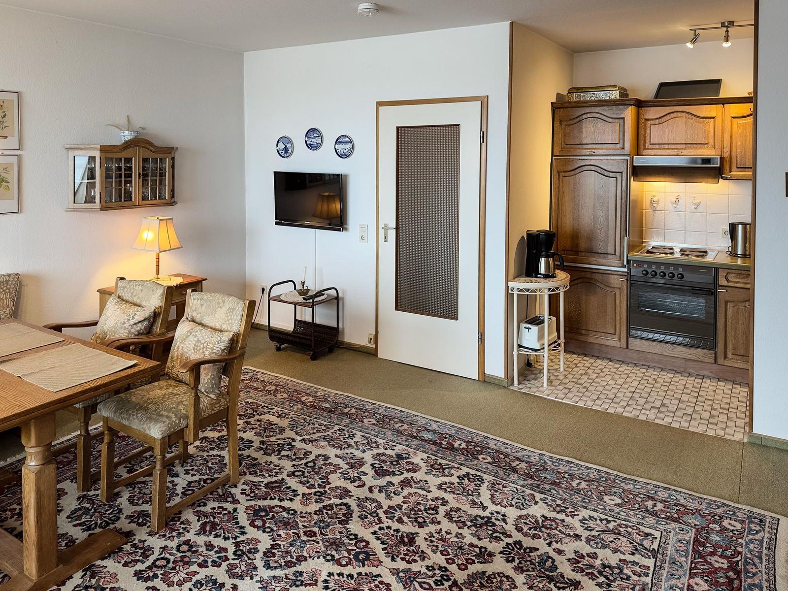Living area with dining space, kitchen, and TV. Wooden furniture and carpeted floor.