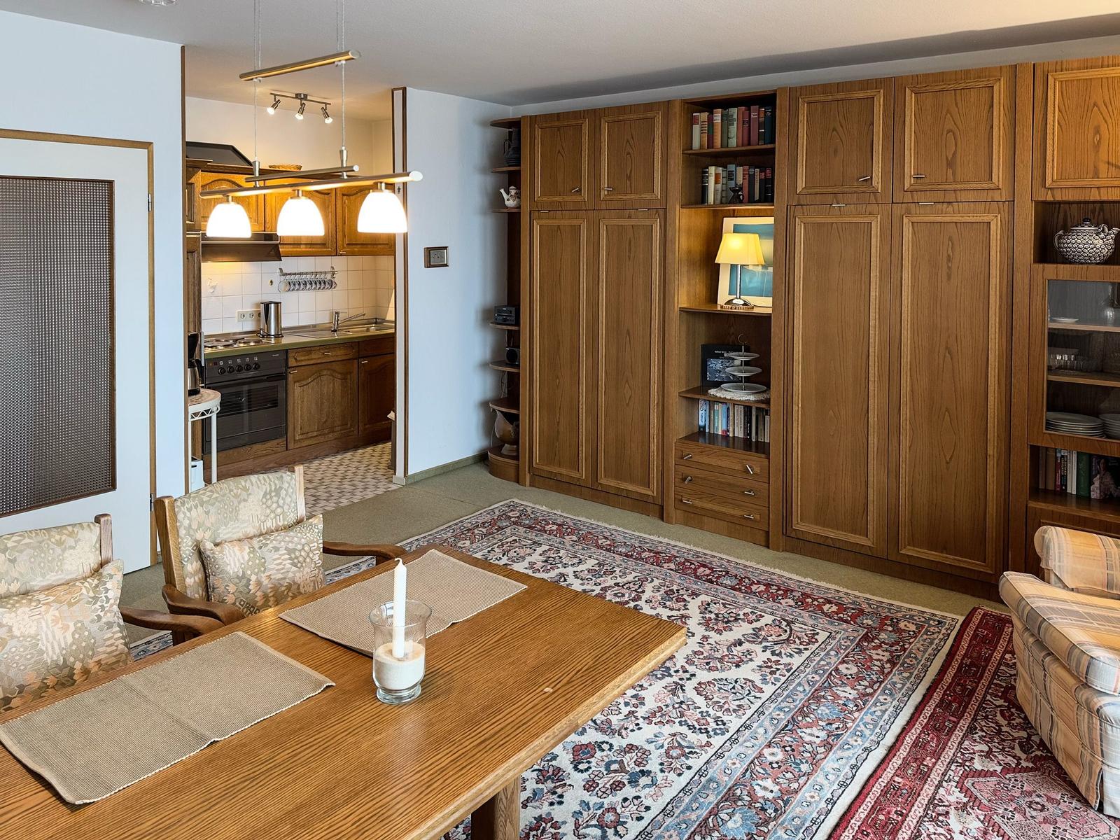 Dining area with wooden table, chairs, and rug. Kitchen and bookshelves are visible.