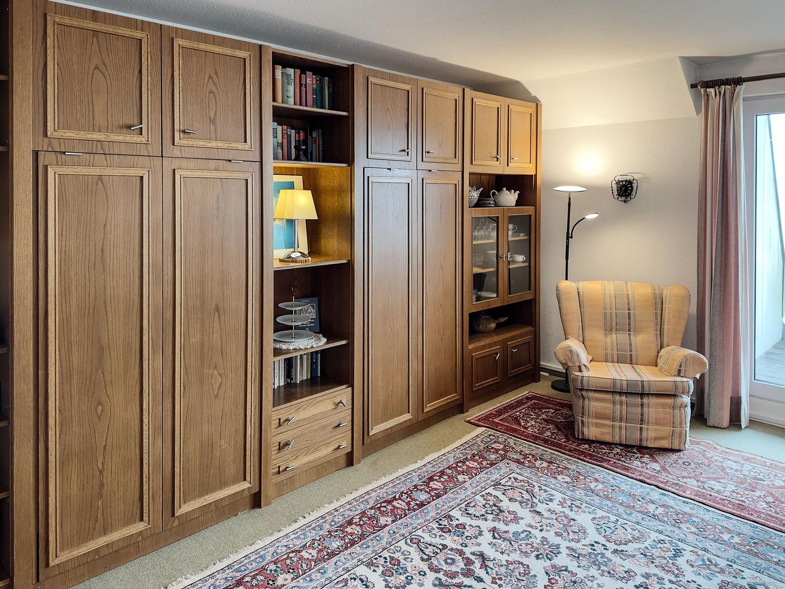 Large wooden wardrobe with shelves and seating. Rug and window are visible.