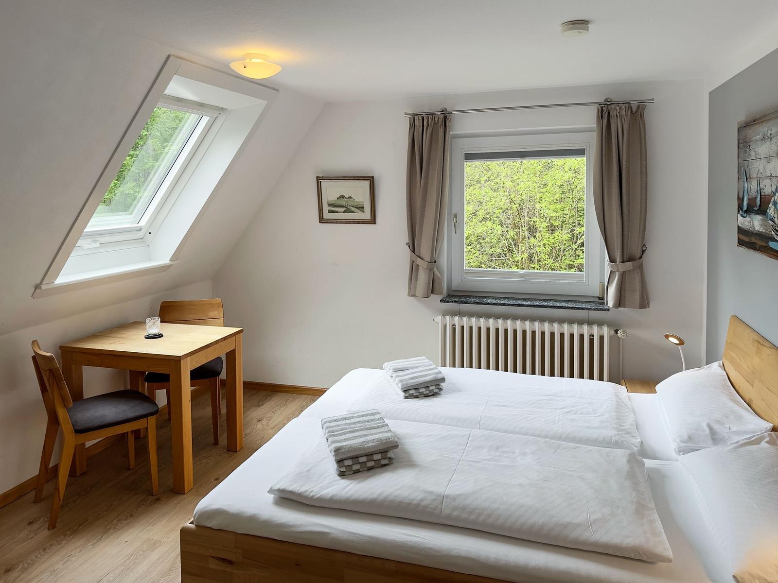 Schlafzimmer mit Doppelbett, Tisch, zwei Stühlen und Fenstern mit Blick auf Grün.