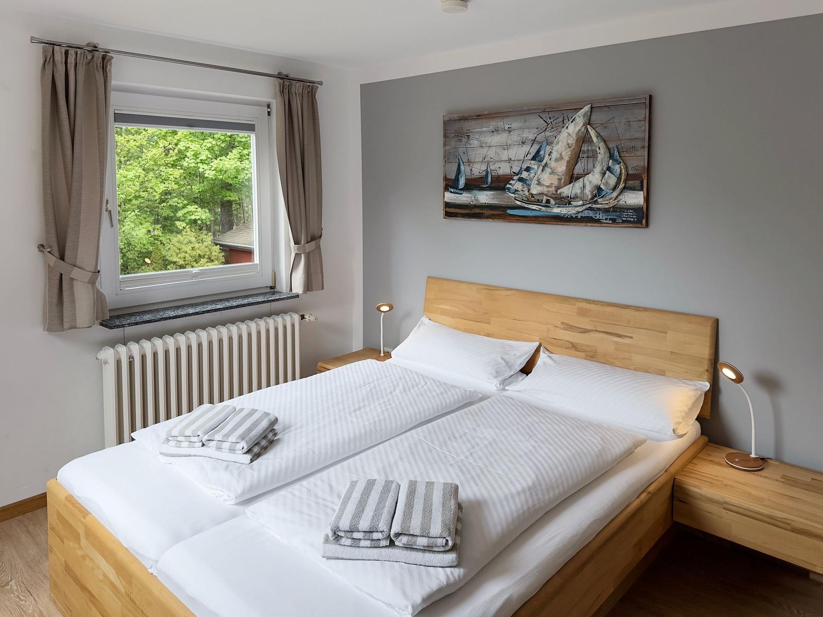 Schlafzimmer mit Holzbett, Fensterblick auf Wald und Wandbild mit Segelboot.