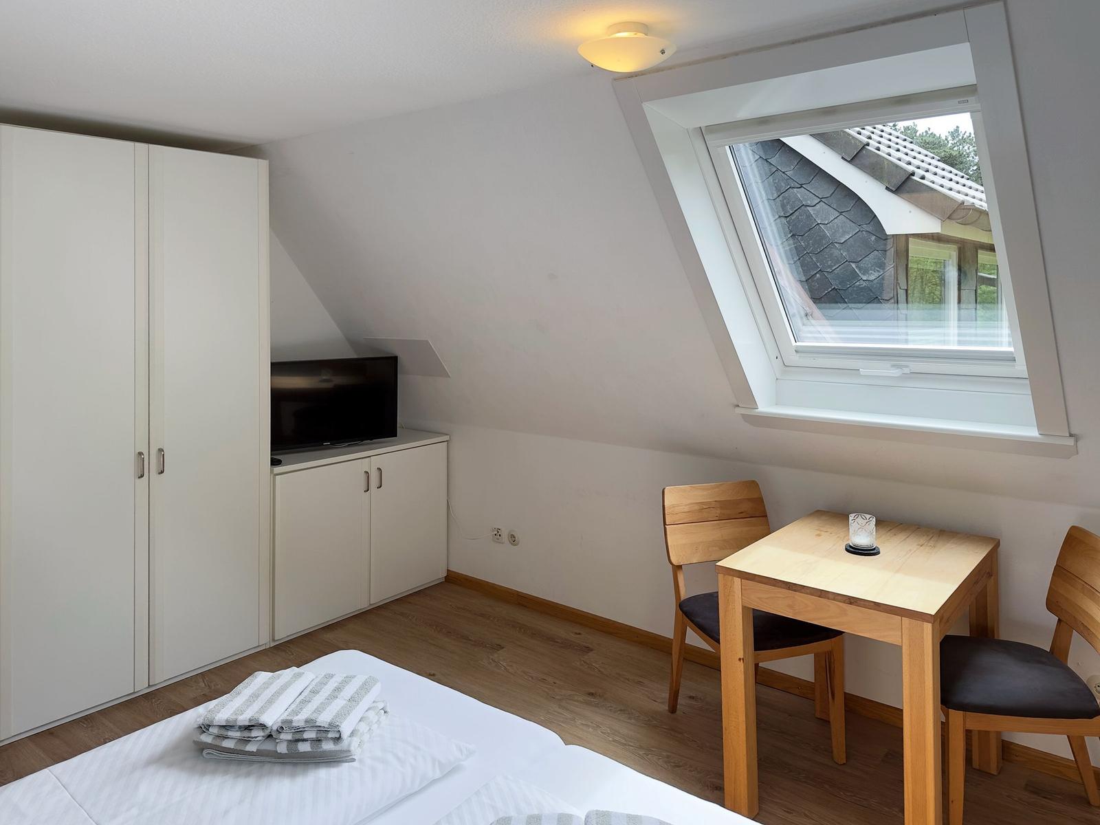 Zimmer mit Dachschrägen, Bett, Kleiderschrank, TV und kleinem Holztisch mit zwei Stühlen.