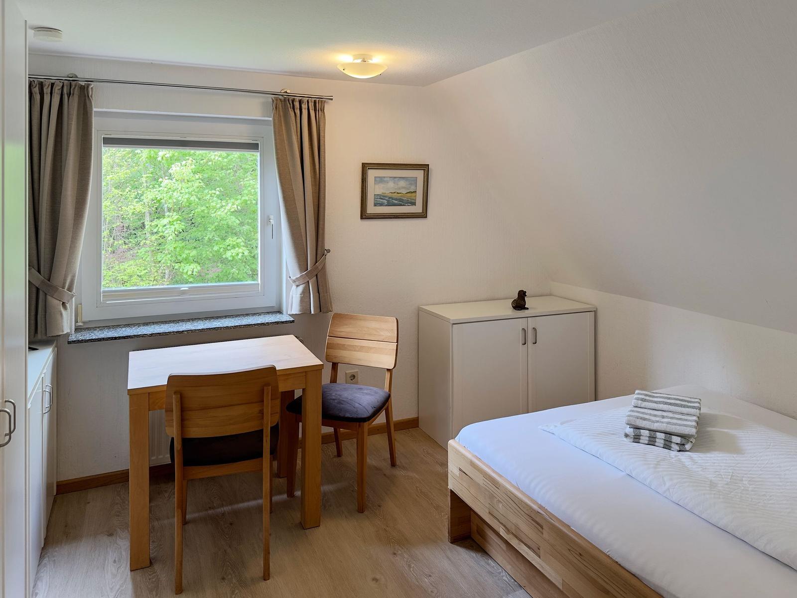 Zimmer mit Bett, Tisch, zwei Stühlen und Fenster mit Blick auf Wald.