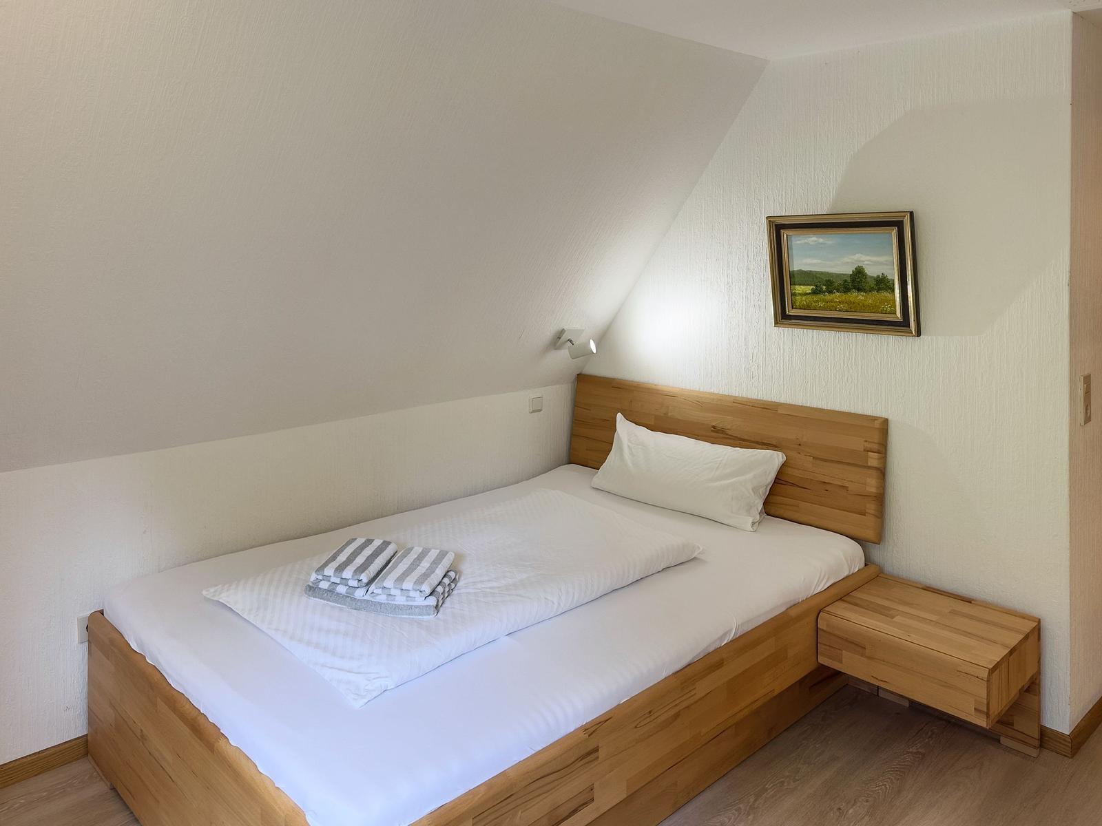 Ein Schlafzimmer mit Holzbett, Nachttisch und Wandbild unter schräger Decke.