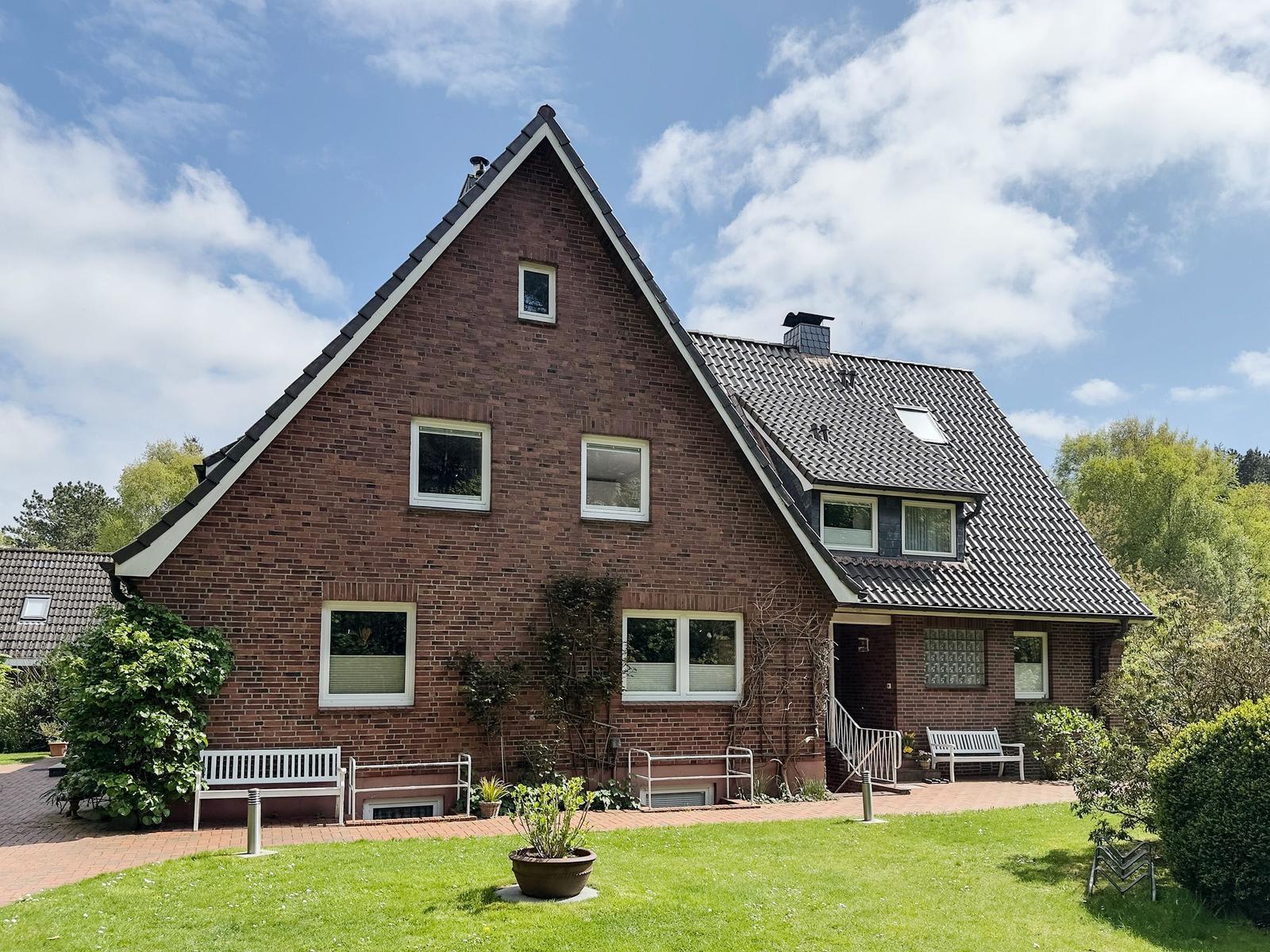 Zweistöckiges Backsteinhaus mit Garten, Terrasse und weißen Bänken unter blauem Himmel.
