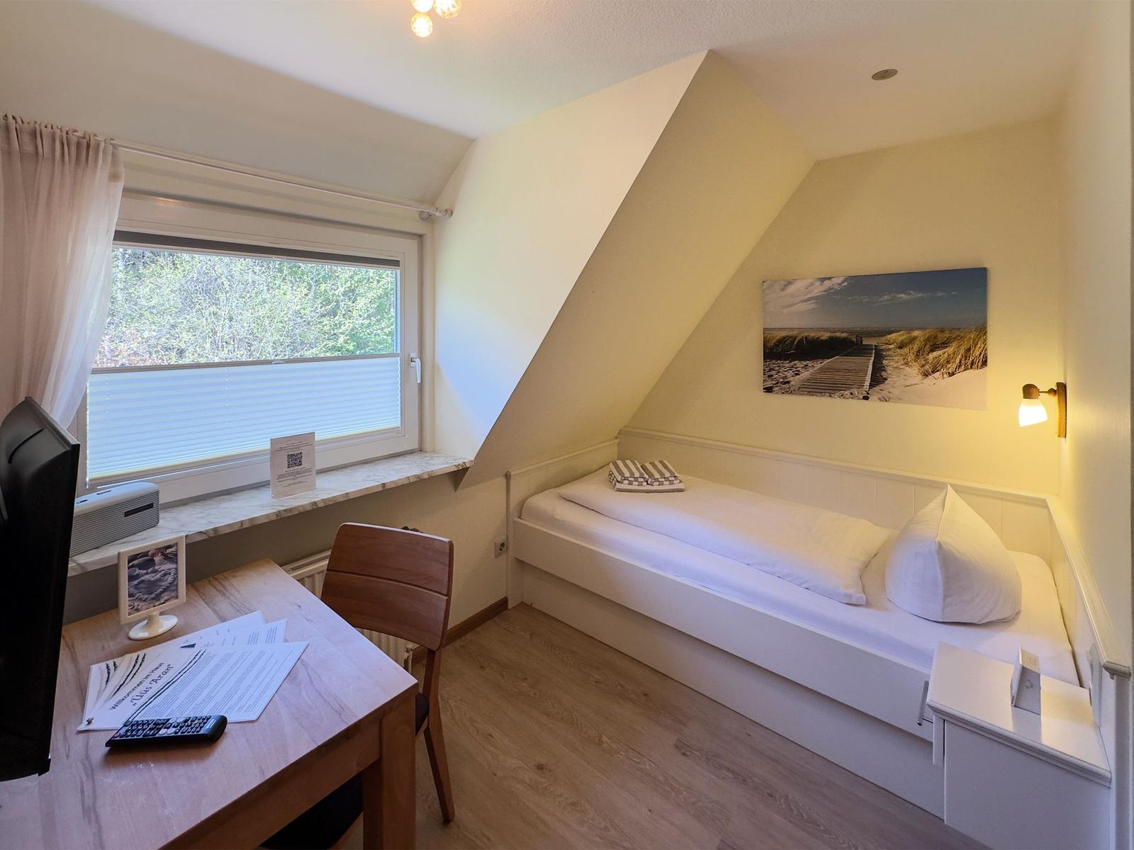Schlafzimmer mit Dachschräge, Bett, Schreibtisch und Fenster mit Blick auf Wald.