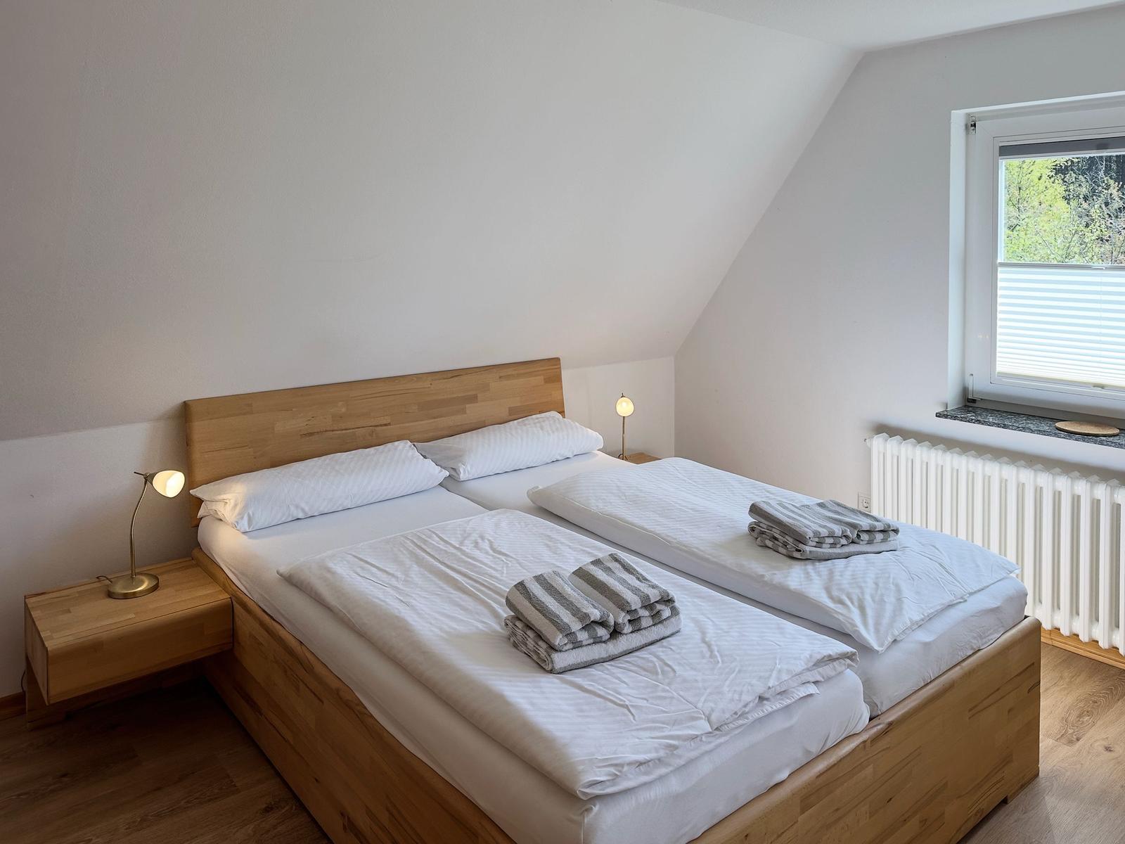 Zimmer mit Doppelbett, Holzbett, Nachttisch und Fenster mit Blick auf Bäume.