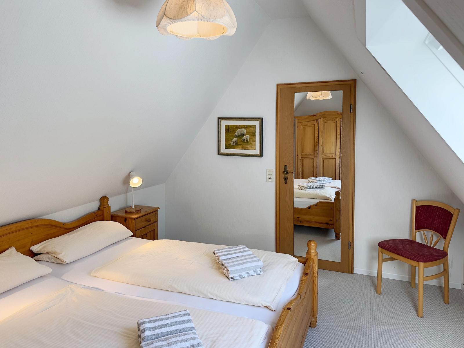 Schlafzimmer mit Holzbett, Nachttisch, Stuhl und Spiegel. Dachschräge und Fenster sichtbar.