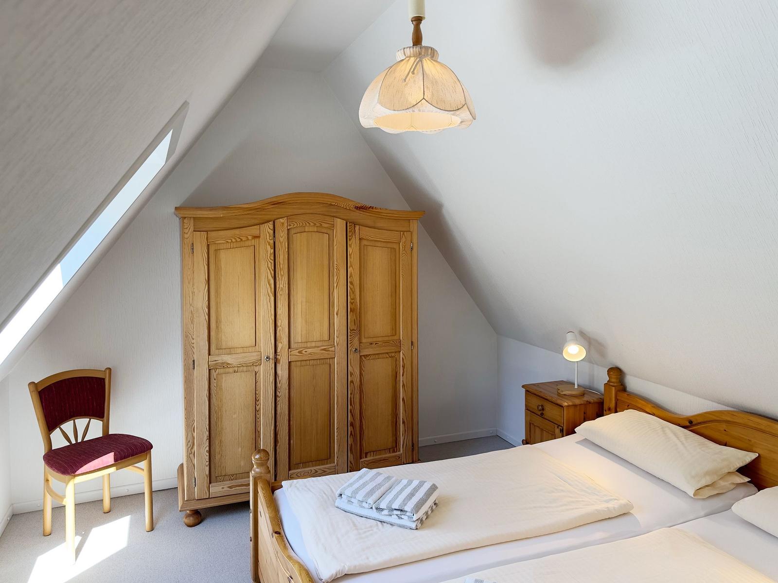 Schlafzimmer mit Dachschrägen, Holzschrank, Bett und Stuhl. Fenster und Lampen sichtbar.