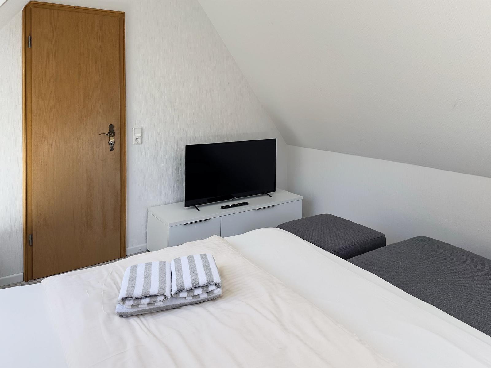Schlafzimmer mit Doppelbett, Fernseher und grauen Sitzkissen.