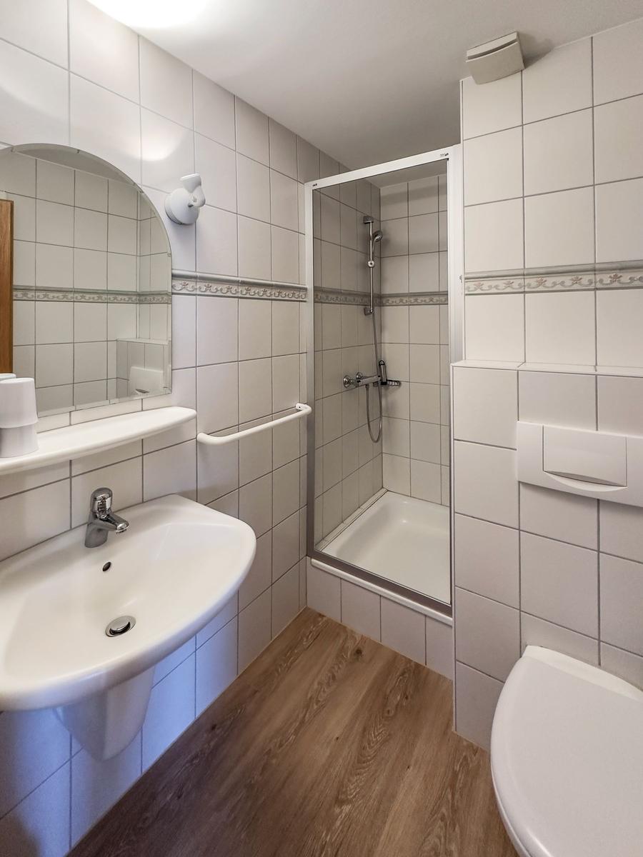 Badezimmer mit Dusche, Waschbecken und Toilette. Fliesenwand und Holzfußboden.