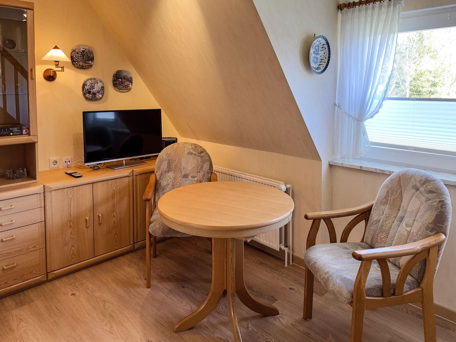 Ein Raum mit Holztisch, zwei Stühlen, Fernseher und Fenster mit Vorhängen.