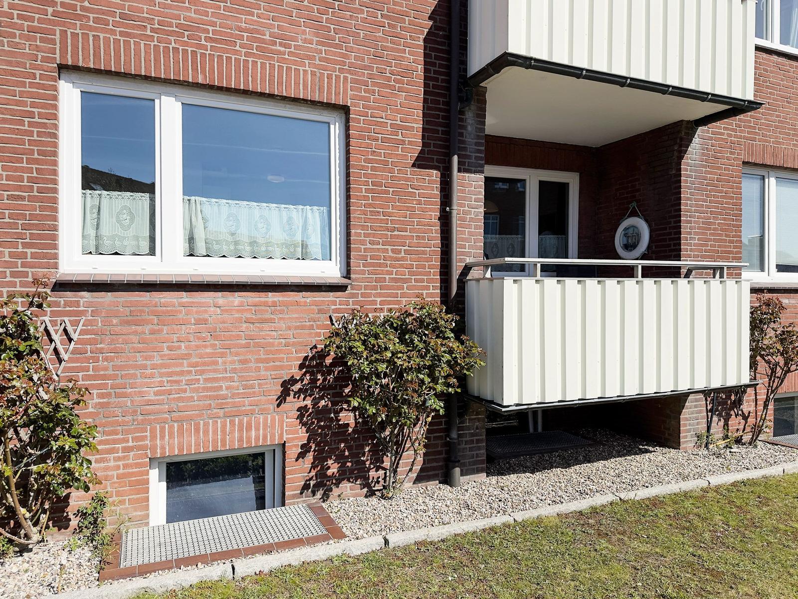 Red brick house with balcony and windows. Grass and shrubs in the front garden.