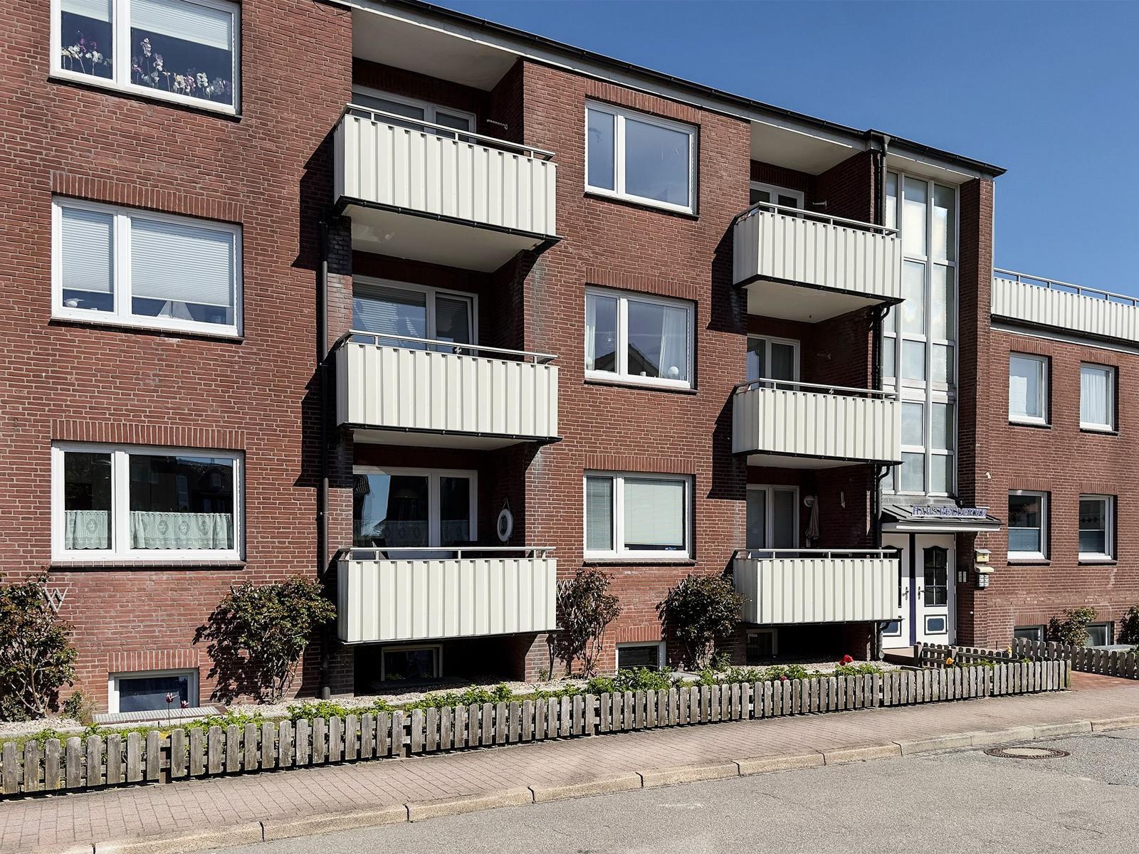 Red brick building with balconies and windows. Front garden with fence.