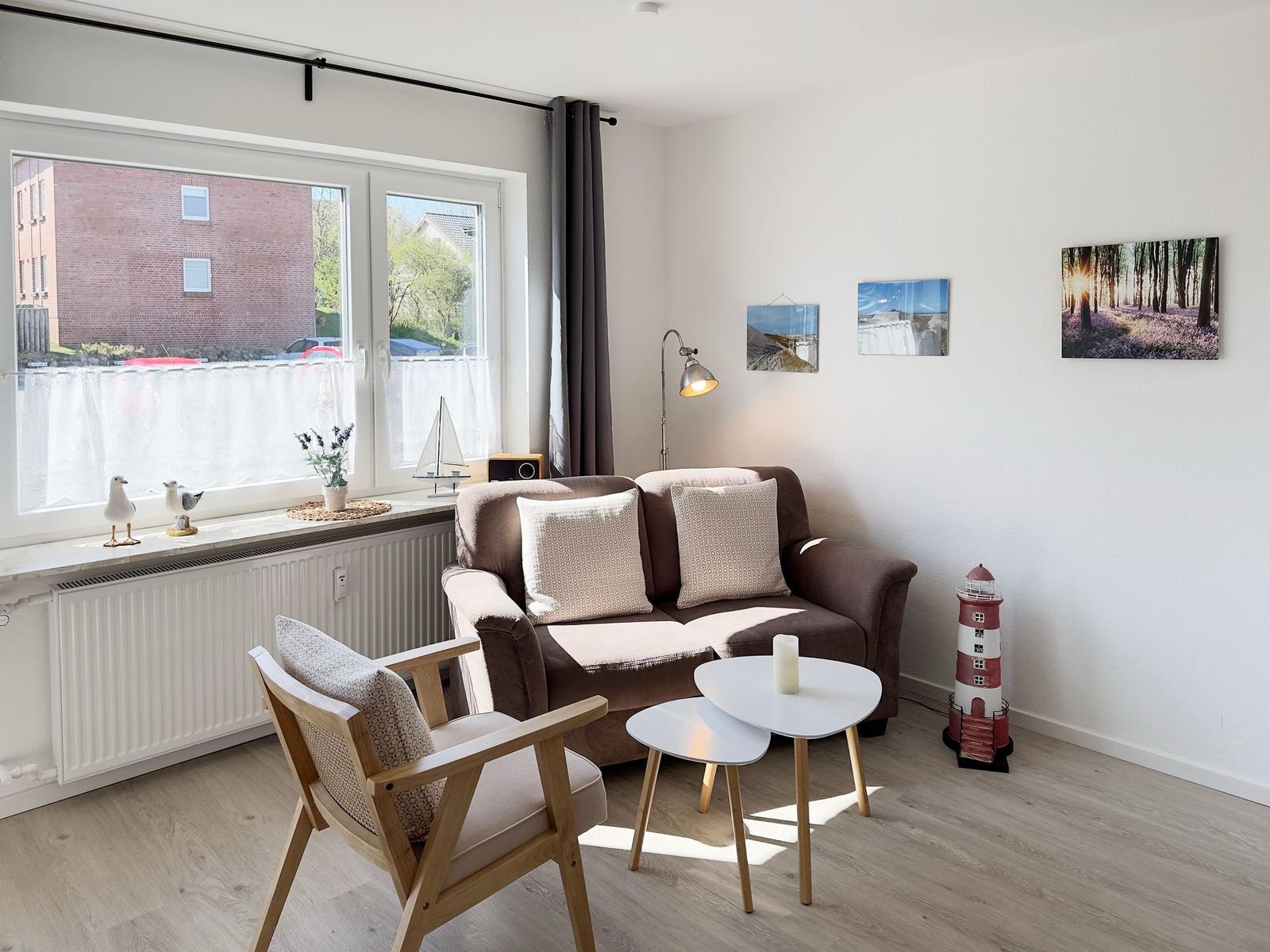 Living room with sofa, chair, and table. Window with view of buildings. Wall art and lighthouse figurine.