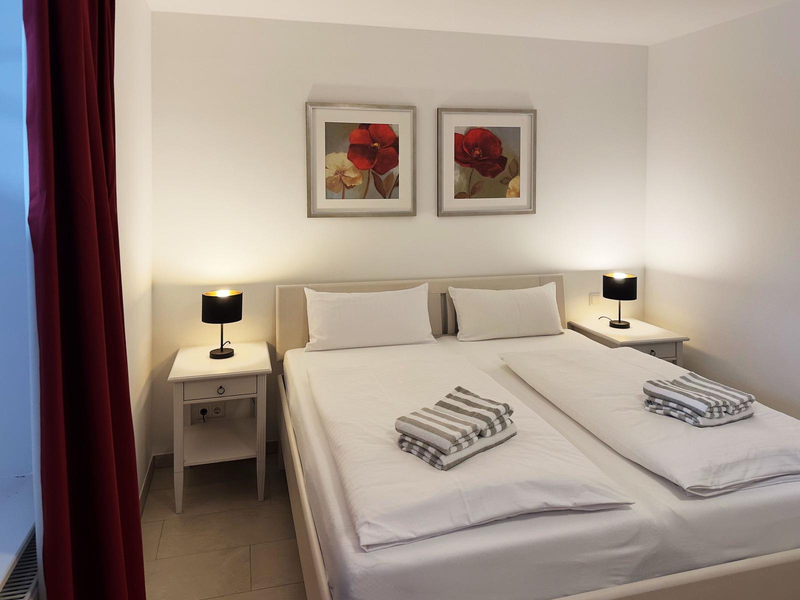Zimmer mit Doppelbett, zwei Nachttischen und zwei Bildern an der Wand.