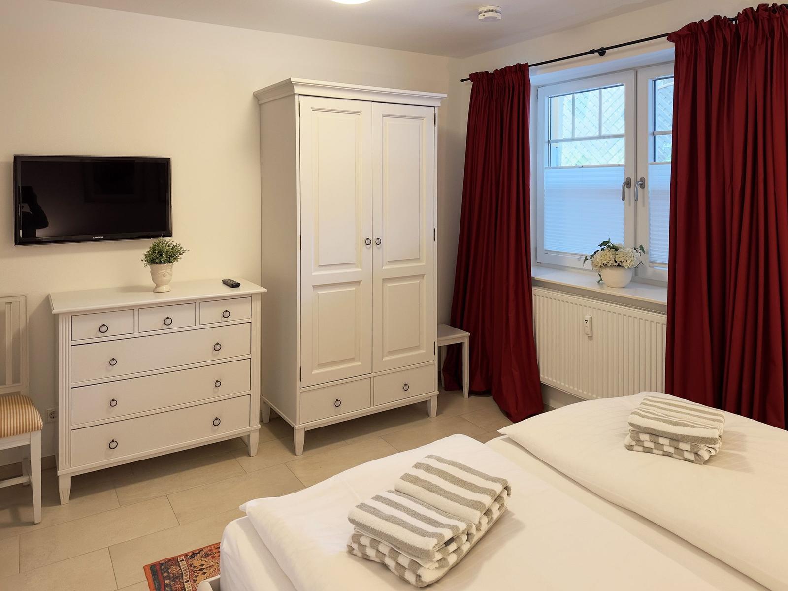 Schlafzimmer mit weißem Schrank, Kommode, Bett und roten Vorhängen.
