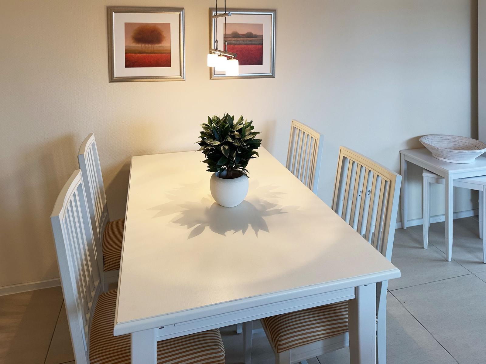 Essbereich mit weißem Tisch, vier Stühlen und Pflanze. An der Wand hängen zwei Bilder.