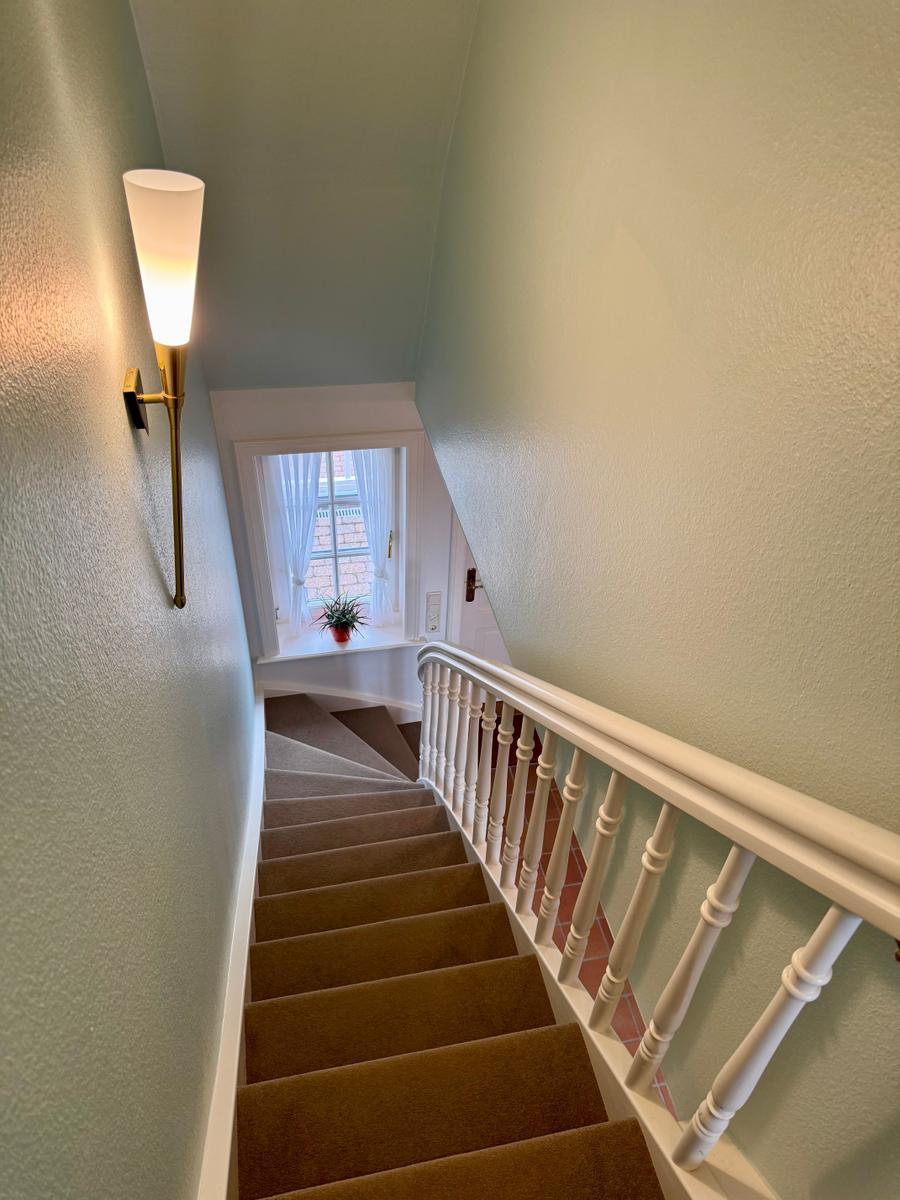Treppe mit Teppich, weißer Handlauf und Wandlampe. Fenster mit Vorhängen im oberen Bereich.