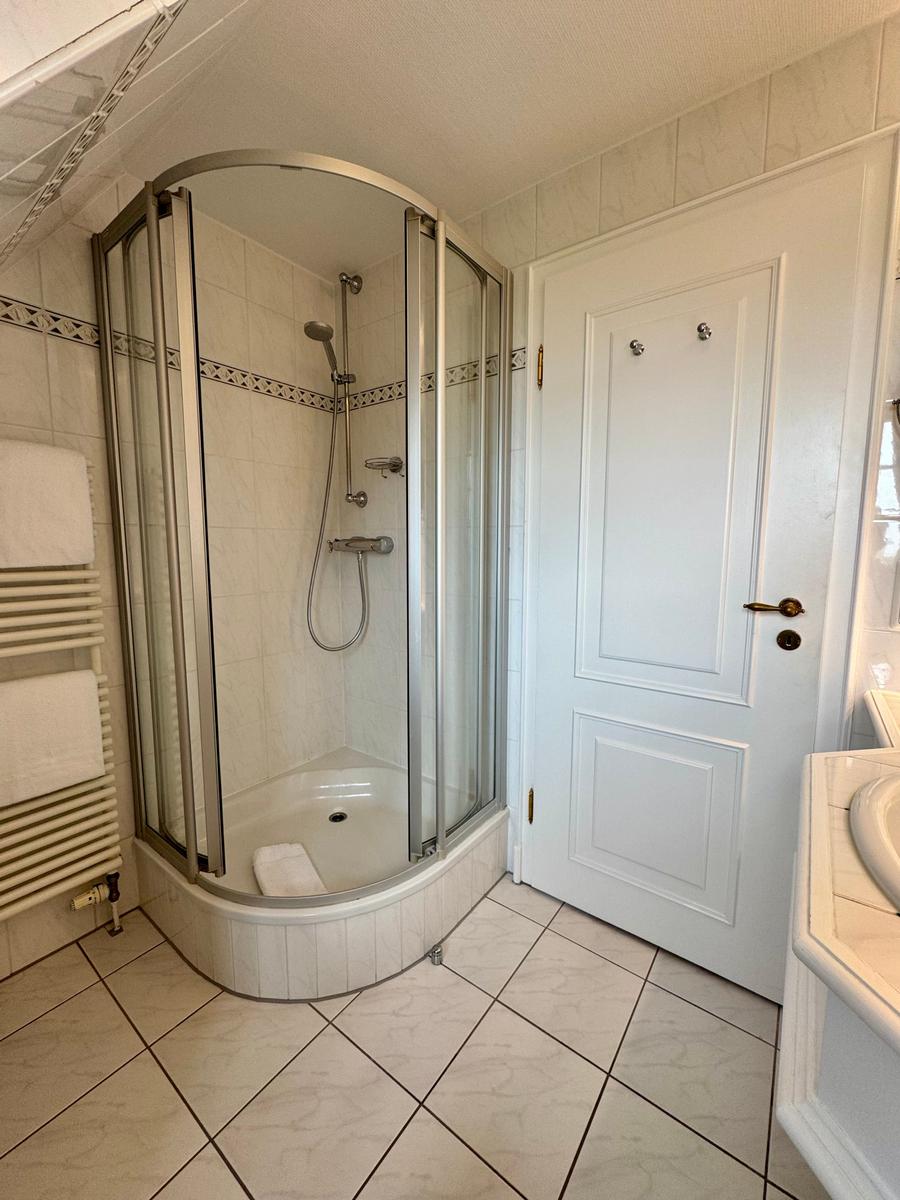 Badezimmer mit Duschkabine, Waschtisch und Heizung. Fliesenboden und weiße Wände.