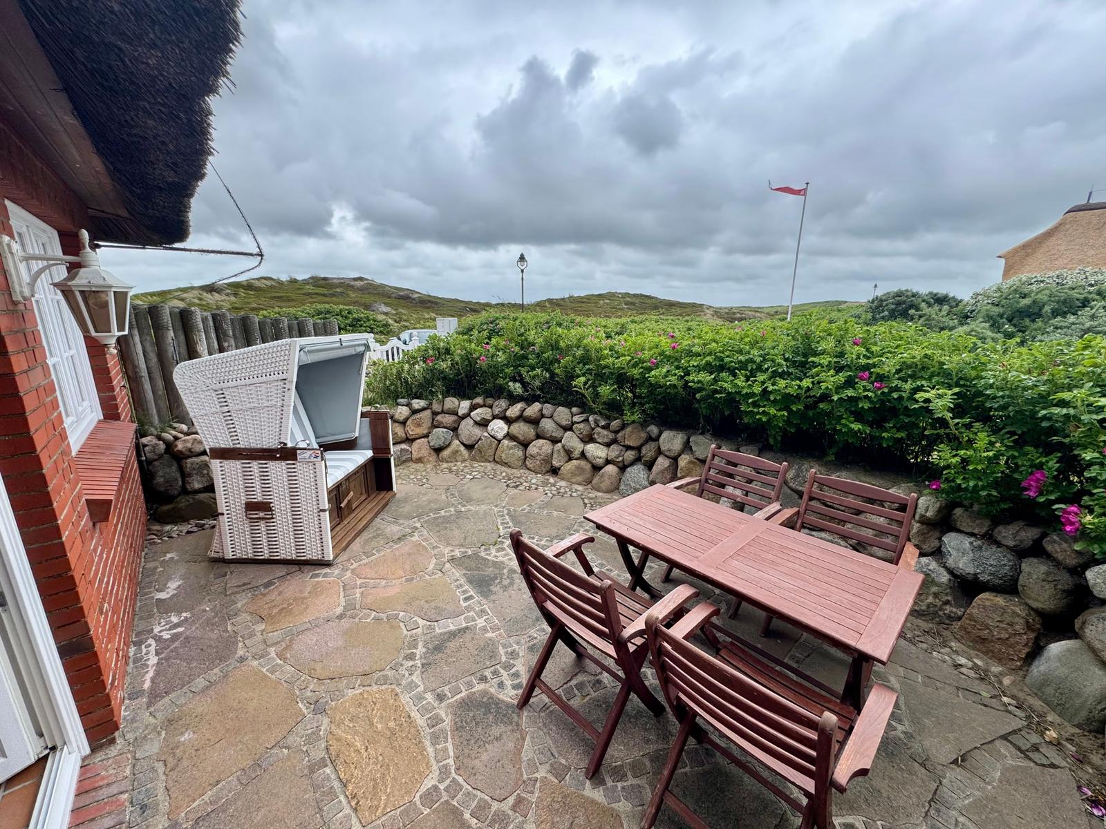 Terrasse mit Tisch, Stühlen und Liege. Steinplatten, Steinmauer, Grünflächen und Flagge.