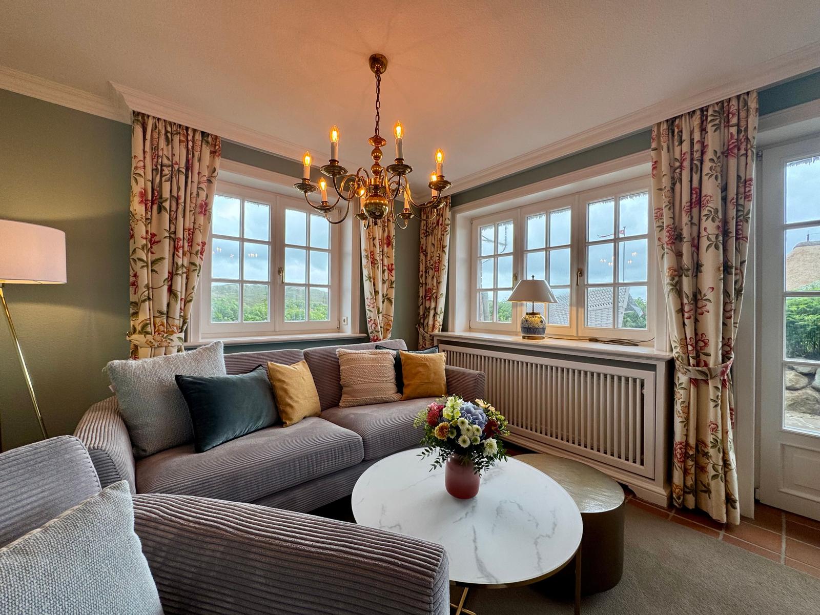 Ein Wohnzimmer mit Sofa, runden Tisch und Fenstern mit floralen Vorhängen.