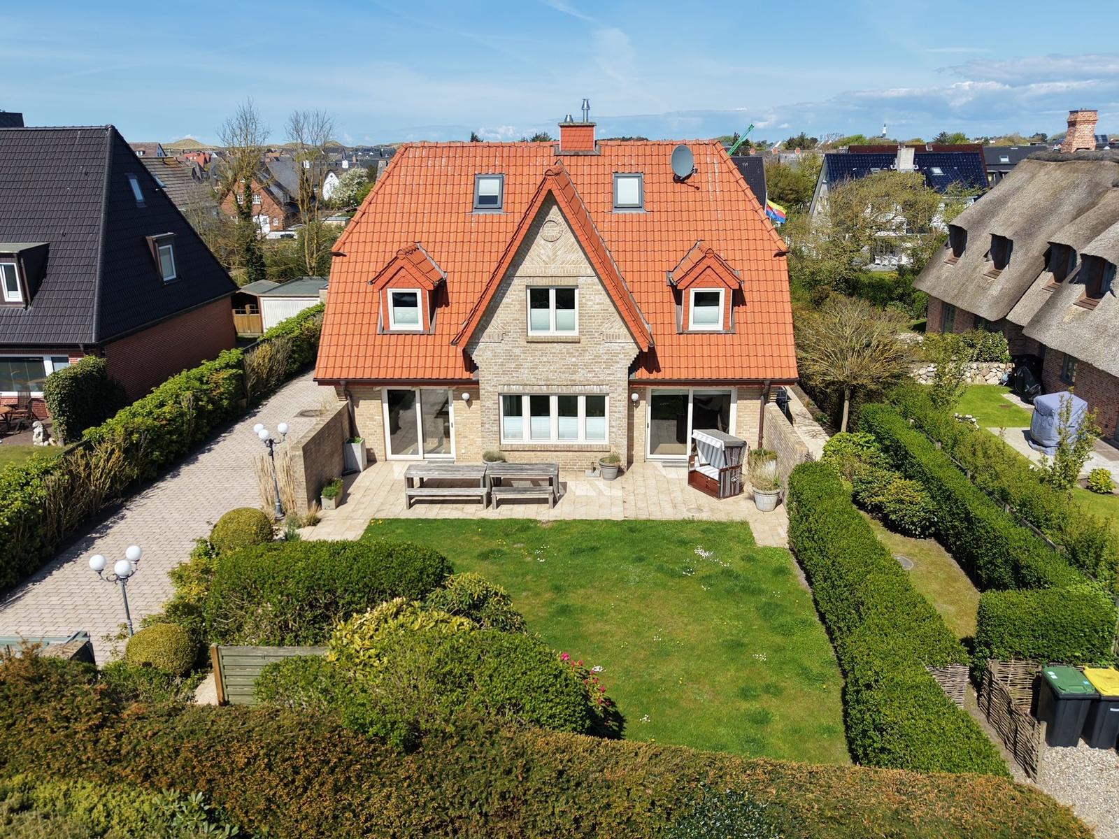 Haus mit rotem Dach, Garten und Terrasse. Umgeben von grüner Vegetation und Nachbarhäusern.