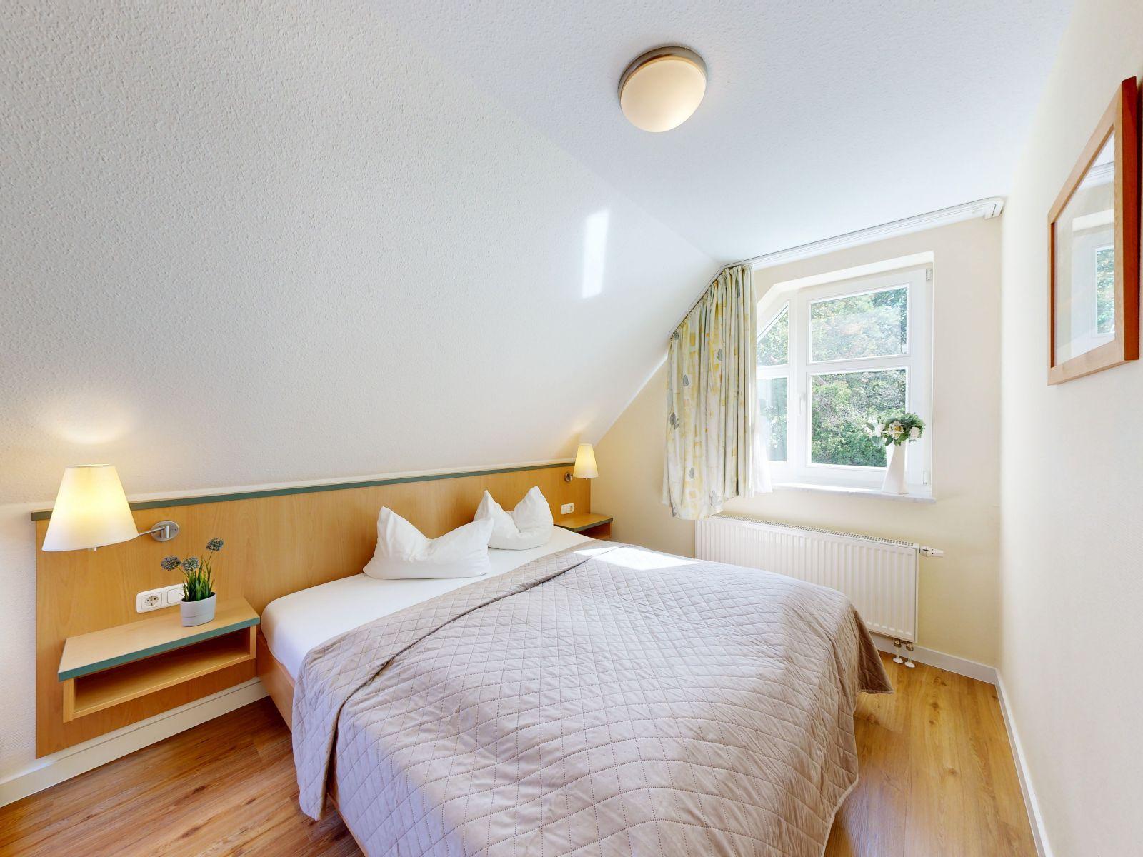 Ein Schlafzimmer mit Dachschrägen, Bett, Nachttisch und Fenster mit Blick auf Bäume.