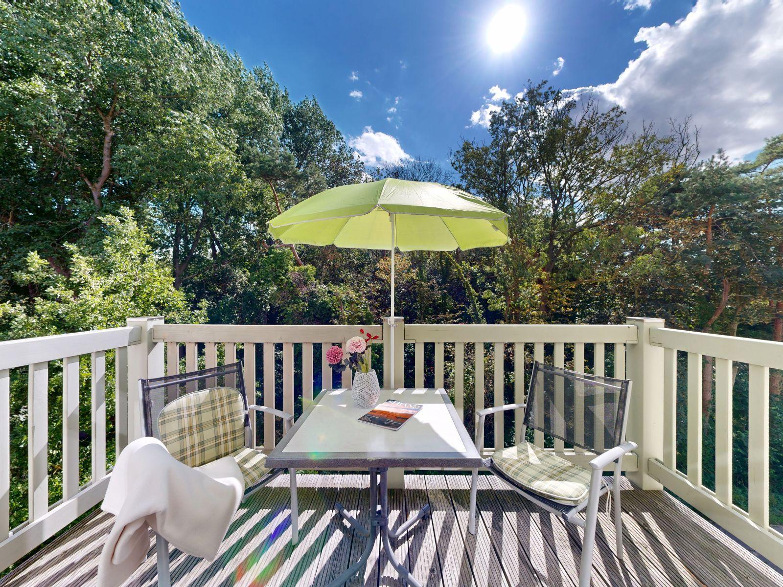 Terrasse mit Tisch, zwei Stühlen und Sonnenschirm. Blick in einen grünen Wald.