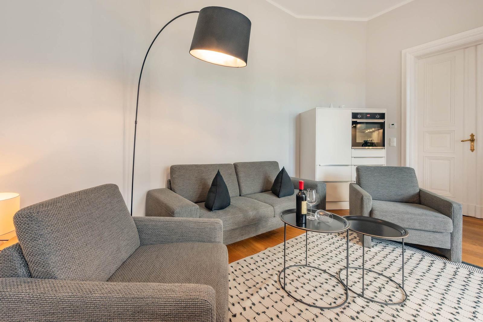 Wohnbereich mit grauem Sofa, zwei Sesseln und einem Tisch mit Wein. Hintergrund: Küchenzeile und Tür.